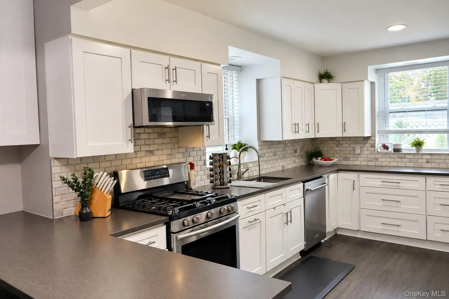 Kitchen featuring white shaker-style cabinetry, stainless steel appliances, dark composite countertops, and a light-toned brick-style tile backsplash Kitchen featuring white shaker-style cabinetry, stainless steel appliances, dark composite countertops, and a light-toned brick-style tile backsplash