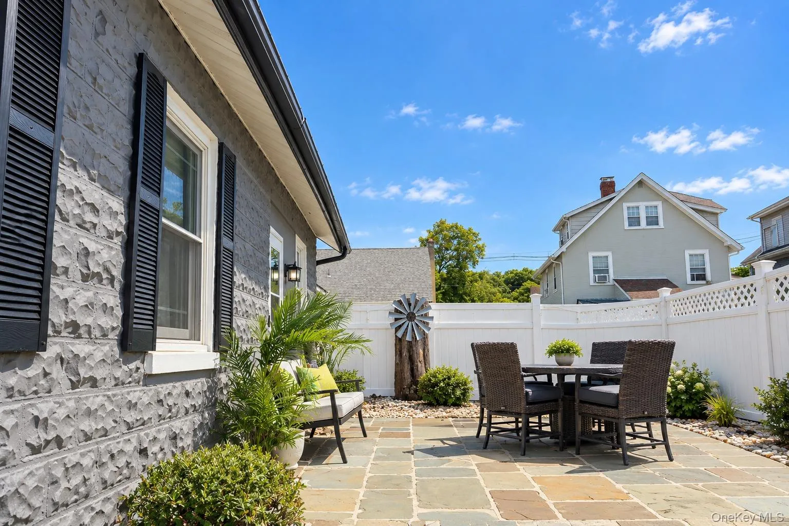 Exterior featuring grey rock-faced masonry siding, dark window shutters, natural stone paver patio, white vinyl privacy fencing, and metal decorative accent Exterior featuring grey rock-faced masonry siding, dark window shutters, natural stone paver patio, white vinyl privacy fencing, and metal decorative accent