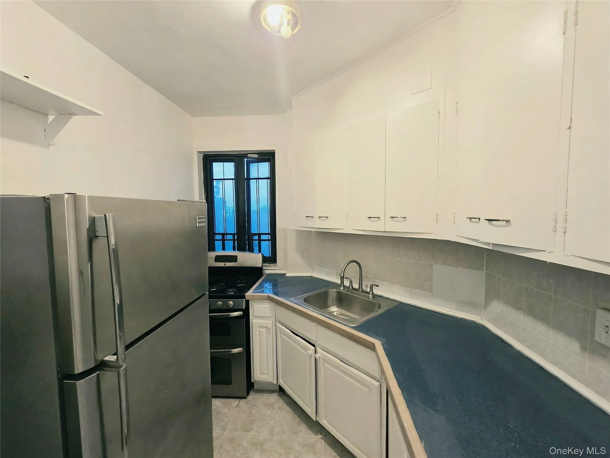 Kitchen featuring white shaker-style cabinetry, stainless steel appliances, dark solid-surface countertops, stainless steel sink with high-arch faucet, and tile flooring Kitchen featuring white shaker-style cabinetry, stainless steel appliances, dark solid-surface countertops, stainless steel sink with high-arch faucet, and tile flooring