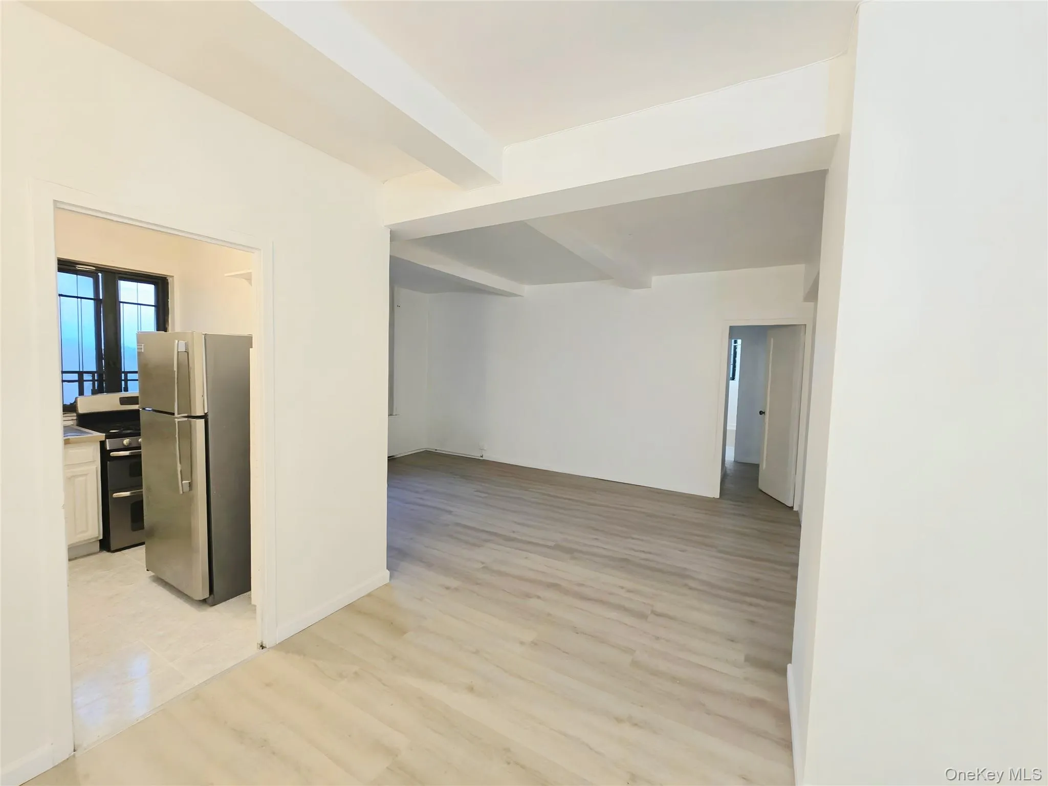 Open living area featuring wood-finish flooring and exposed structural ceiling beams, with a kitchen containing a stainless steel refrigerator and range Open living area featuring wood-finish flooring and exposed structural ceiling beams, with a kitchen containing a stainless steel refrigerator and range