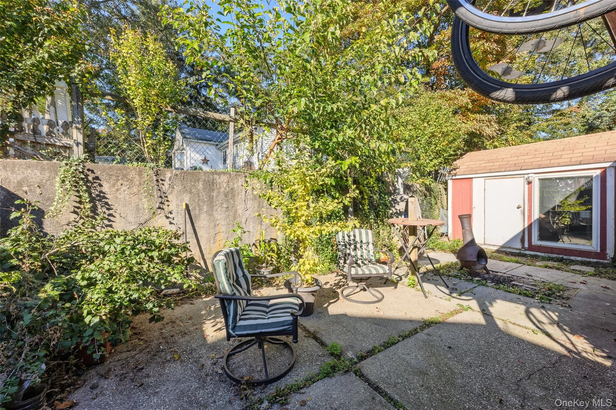 Concrete patio featuring a masonry retaining wall, detached storage structure with asphalt shingle roofing, and established perimeter vegetation Concrete patio featuring a masonry retaining wall, detached storage structure with asphalt shingle roofing, and established perimeter vegetation