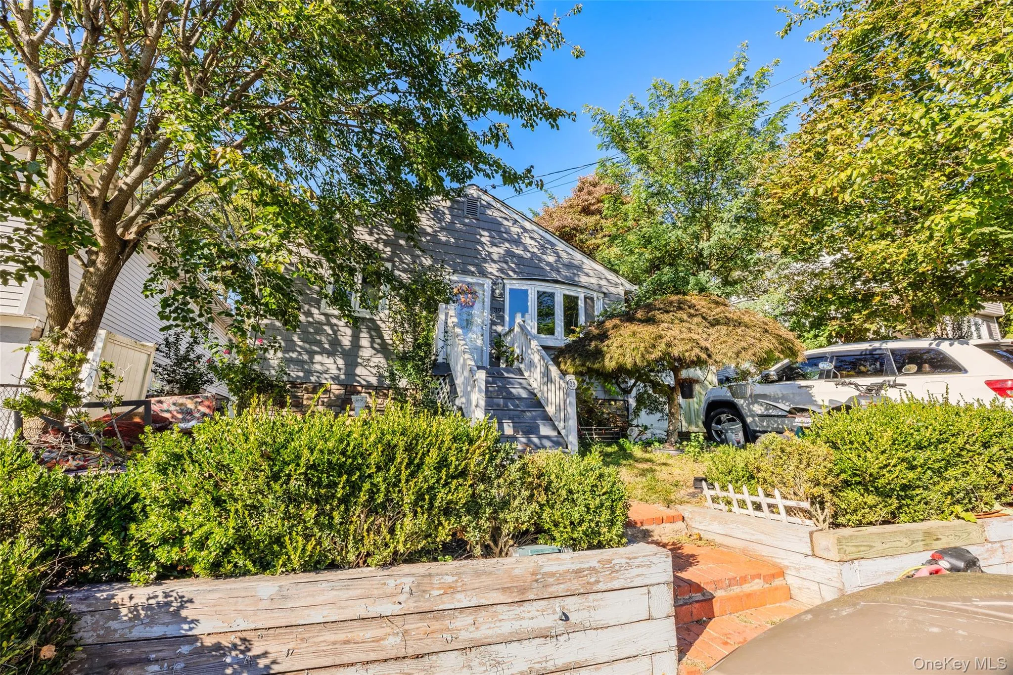 Exterior featuring gray siding, a prominent bay window, timber retaining walls, brick stair entry, and mature landscaping Exterior featuring gray siding, a prominent bay window, timber retaining walls, brick stair entry, and mature landscaping