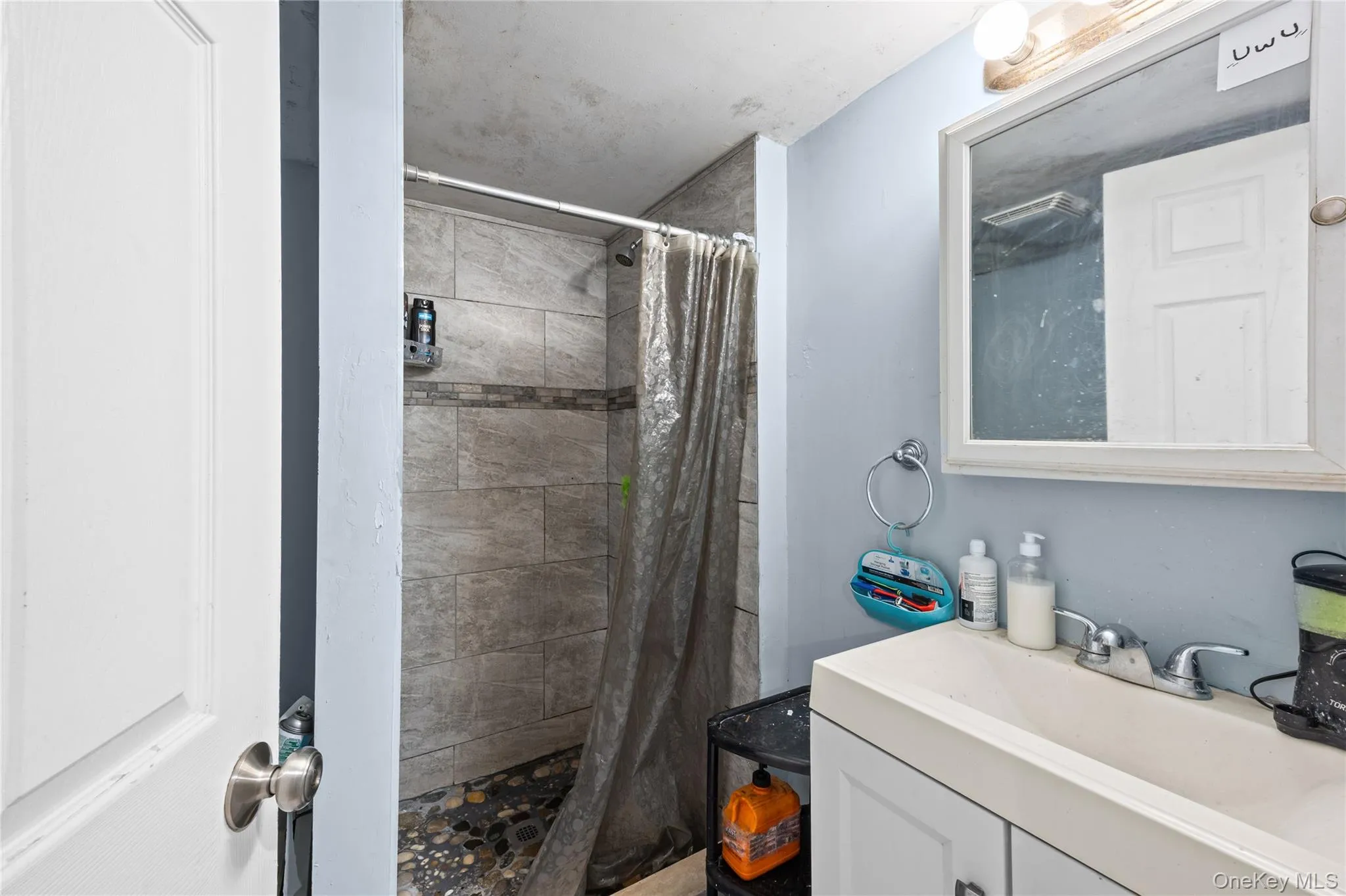 Bathroom featuring a shower stall with stone-finish tile surround, a pebble-style floor, a white vanity with integrated sink, and a wall-mounted medicine cabinet with a mirror Bathroom featuring a shower stall with stone-finish tile surround, a pebble-style floor, a white vanity with integrated sink, and a wall-mounted medicine cabinet with a mirror