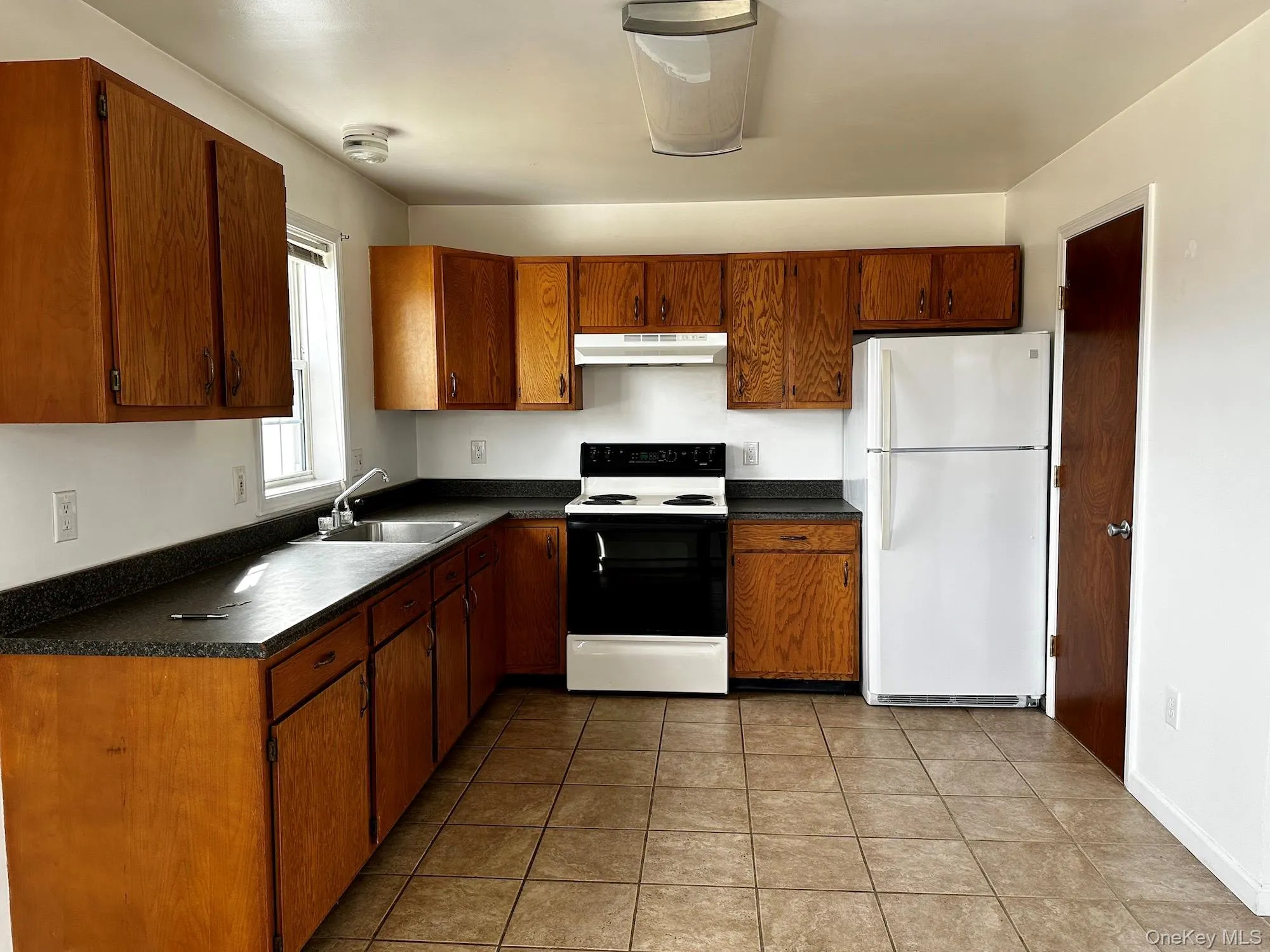 Kitchen with freestanding refrigerator, electric stove, dark countertops, brown cabinetry, and light tile patterned floors Kitchen with freestanding refrigerator, electric stove, dark countertops, brown cabinetry, and light tile patterned floors