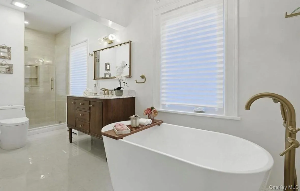 Freestanding soaking tub with a floor-mounted brass faucet, wood-finish vanity with stone countertop, and a walk-in glass-enclosed shower featuring a recessed tile niche Freestanding soaking tub with a floor-mounted brass faucet, wood-finish vanity with stone countertop, and a walk-in glass-enclosed shower featuring a recessed tile niche