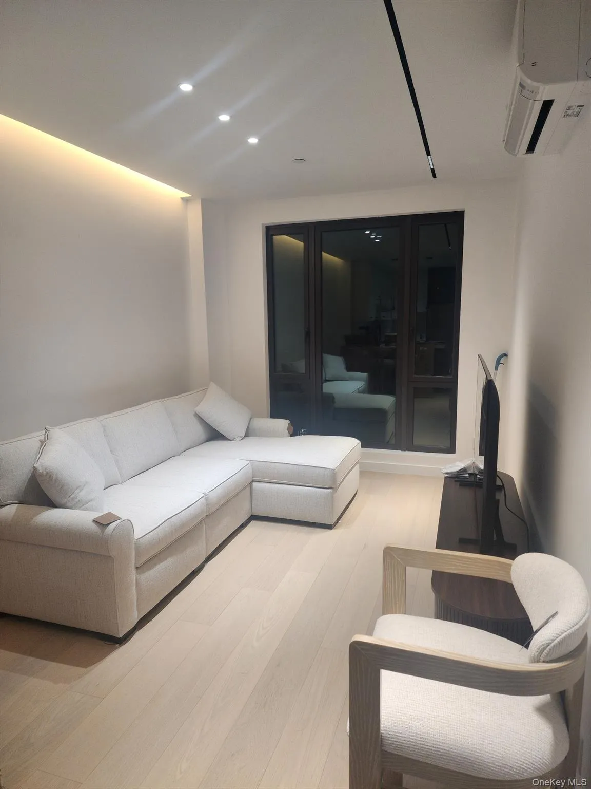 Light-toned wood-finish flooring, recessed ceiling lighting, cove accent lighting, and large dark-framed windows with an integrated wall-mounted climate control unit Light-toned wood-finish flooring, recessed ceiling lighting, cove accent lighting, and large dark-framed windows with an integrated wall-mounted climate control unit