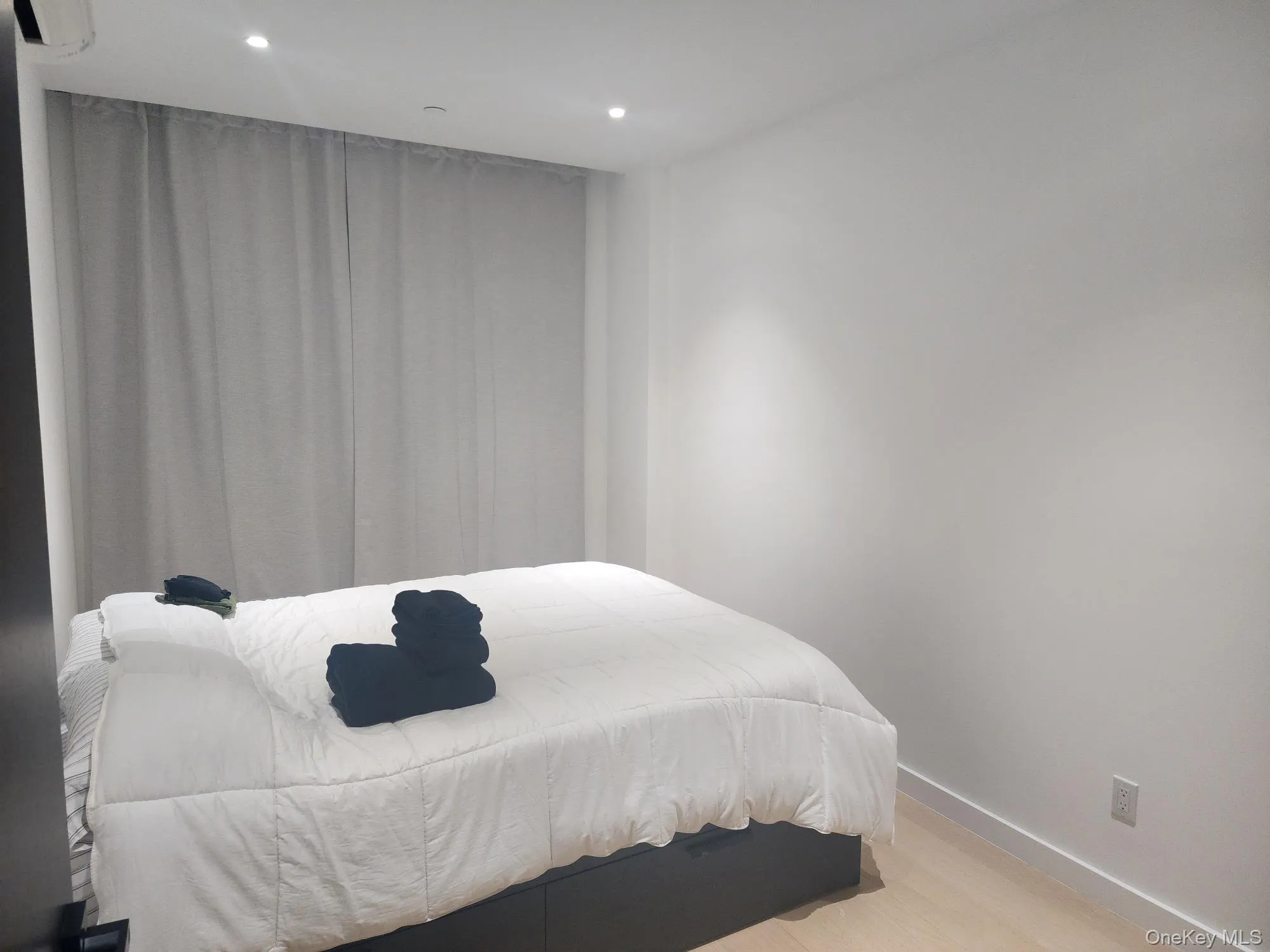 Room featuring neutral walls, recessed ceiling lighting, wood-finish flooring, and a wall-mounted climate control unit Room featuring neutral walls, recessed ceiling lighting, wood-finish flooring, and a wall-mounted climate control unit