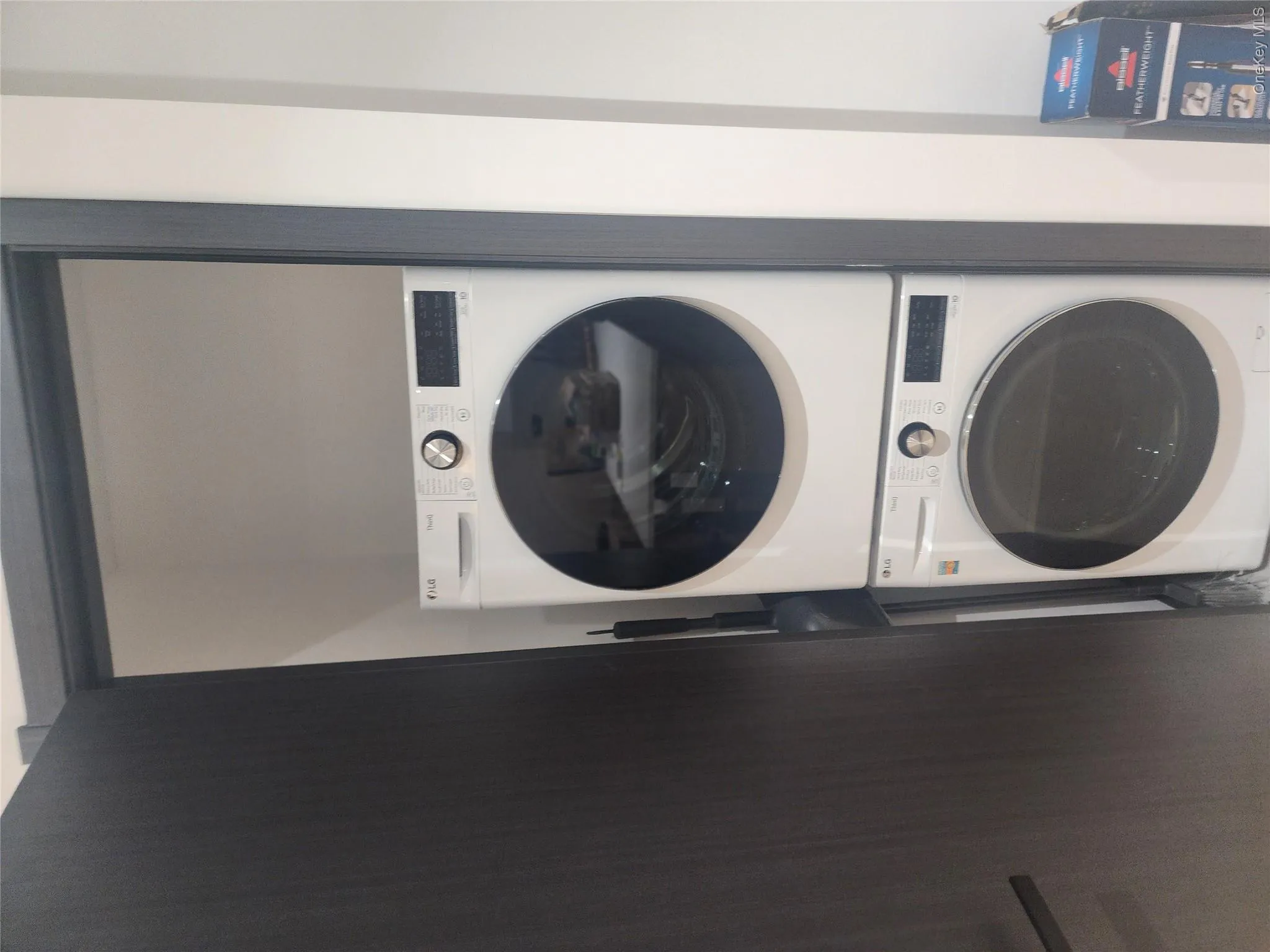 Stacked white washer and dryer unit housed within a dedicated closet featuring a dark wood-finish door and matching door frame Stacked white washer and dryer unit housed within a dedicated closet featuring a dark wood-finish door and matching door frame