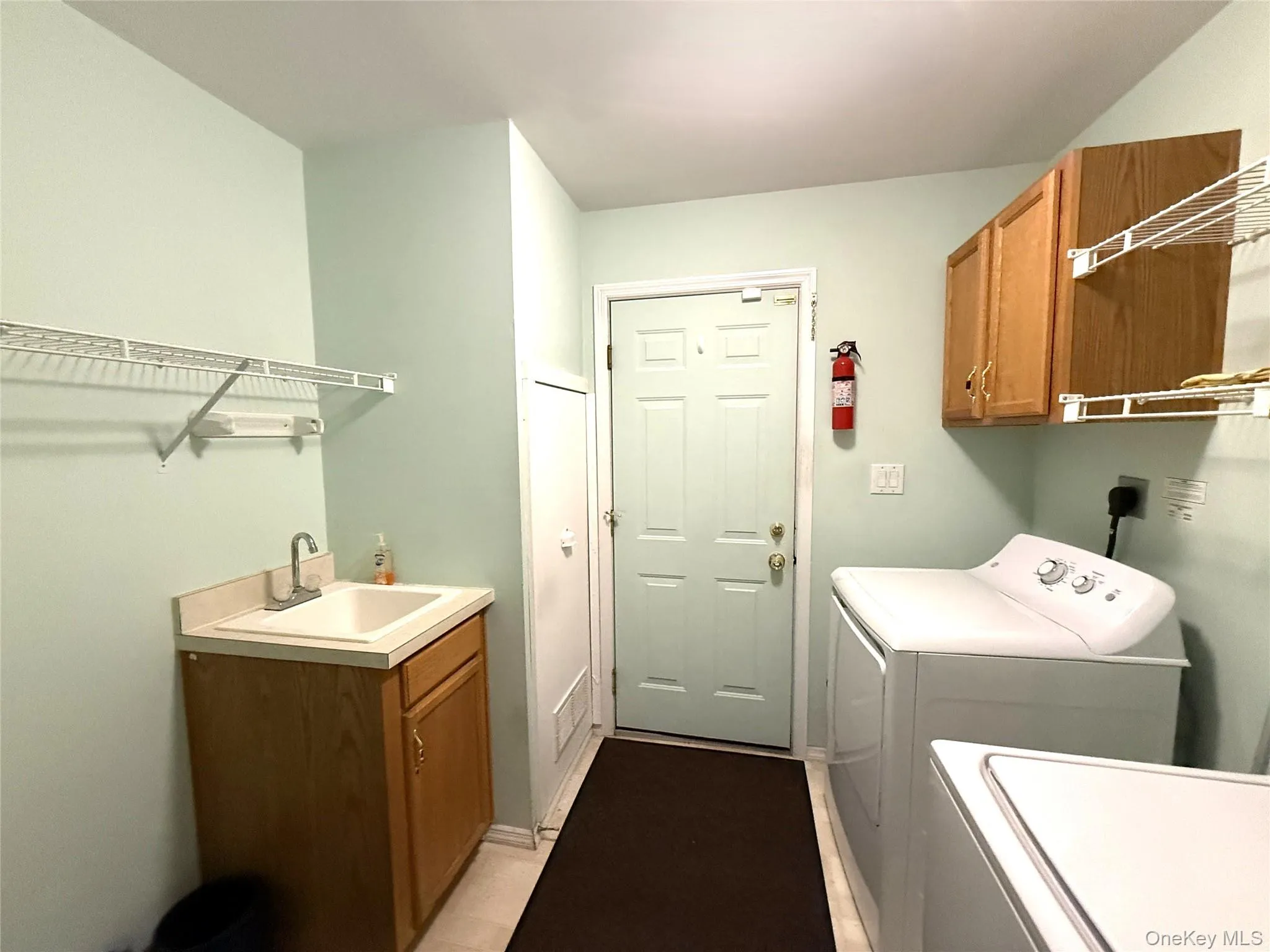 Laundry room featuring a built-in sink with cabinetry, overhead wooden wall cabinets, wire shelving, and a 6 panel door to the garage. Laundry room featuring a built-in sink with cabinetry, overhead wooden wall cabinets, wire shelving, and a 6 panel door to the garage.