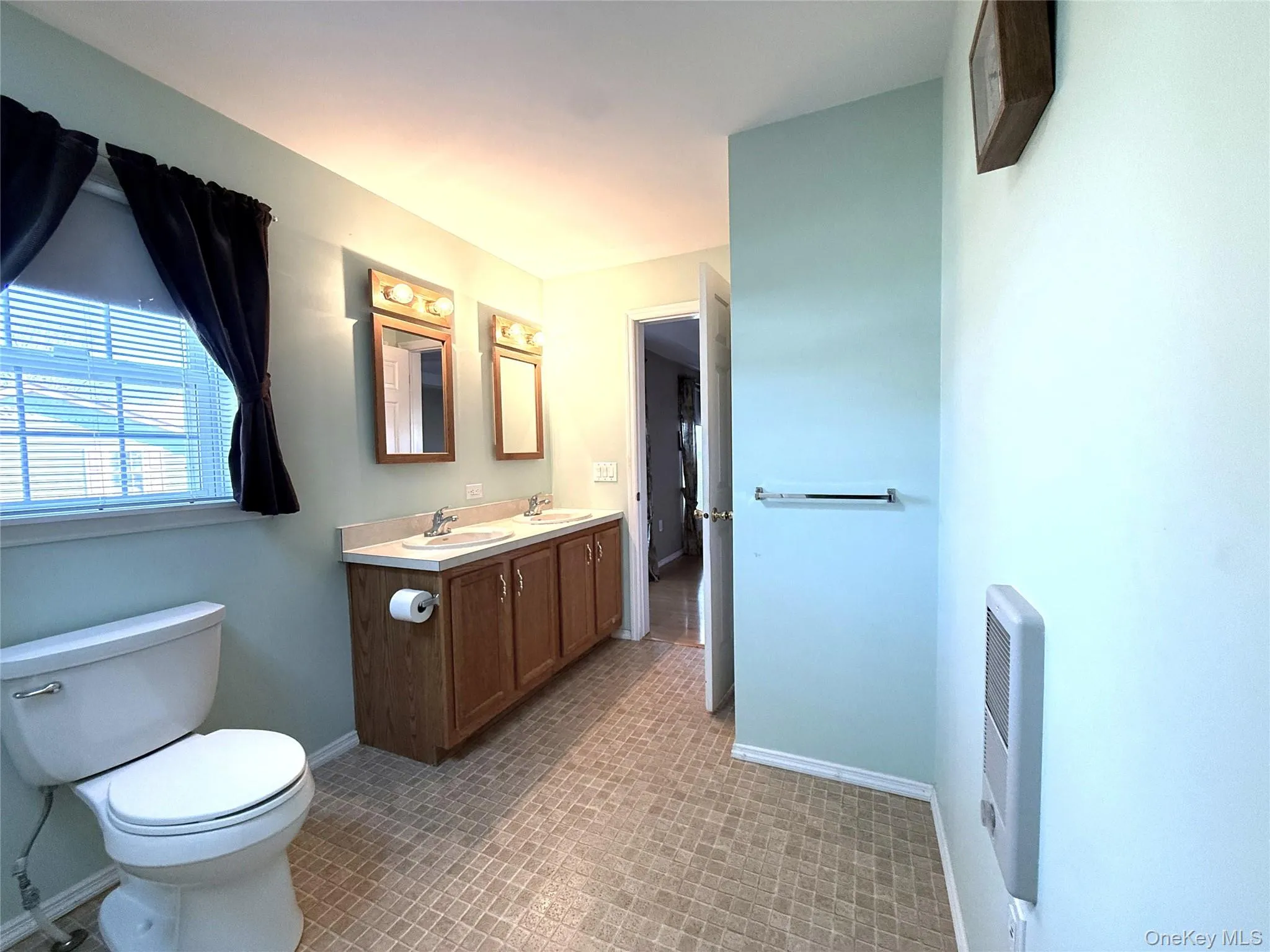 Bathroom featuring a wood-tone dual-sink vanity, solid surface countertop, two wall-mounted mirrors with integrated lighting, and checkered-pattern flooring Bathroom featuring a wood-tone dual-sink vanity, solid surface countertop, two wall-mounted mirrors with integrated lighting, and checkered-pattern flooring