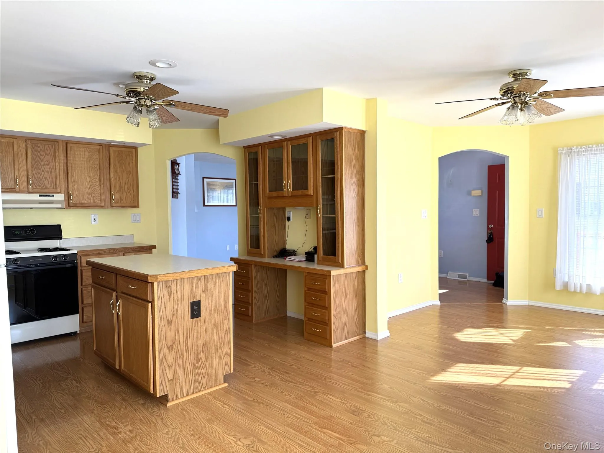 Kitchen featuring wood-finish cabinetry, a central island, built-in desk workstation with overhead glass-front cabinets, wood-finish flooring, and dual ceiling fans Kitchen featuring wood-finish cabinetry, a central island, built-in desk workstation with overhead glass-front cabinets, wood-finish flooring, and dual ceiling fans