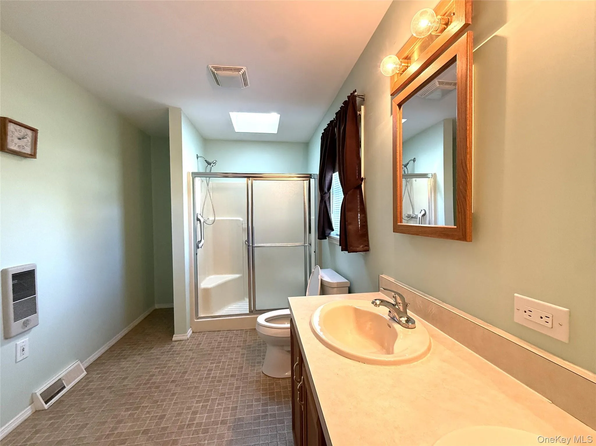 Bathroom featuring a vanity with integrated dual sink, skylight, shower enclosure with sliding glass doors, wall-mounted wood-frame mirror, and wall-mounted light fixture Bathroom featuring a vanity with integrated dual sink, skylight, shower enclosure with sliding glass doors, wall-mounted wood-frame mirror, and wall-mounted light fixture