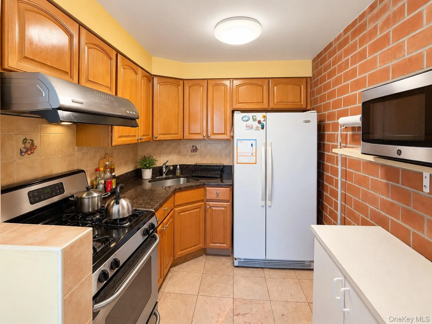 Kitchen featuring wood cabinetry, stone countertops, exposed brick wall, tile flooring, and integrated stainless steel appliances Kitchen featuring wood cabinetry, stone countertops, exposed brick wall, tile flooring, and integrated stainless steel appliances