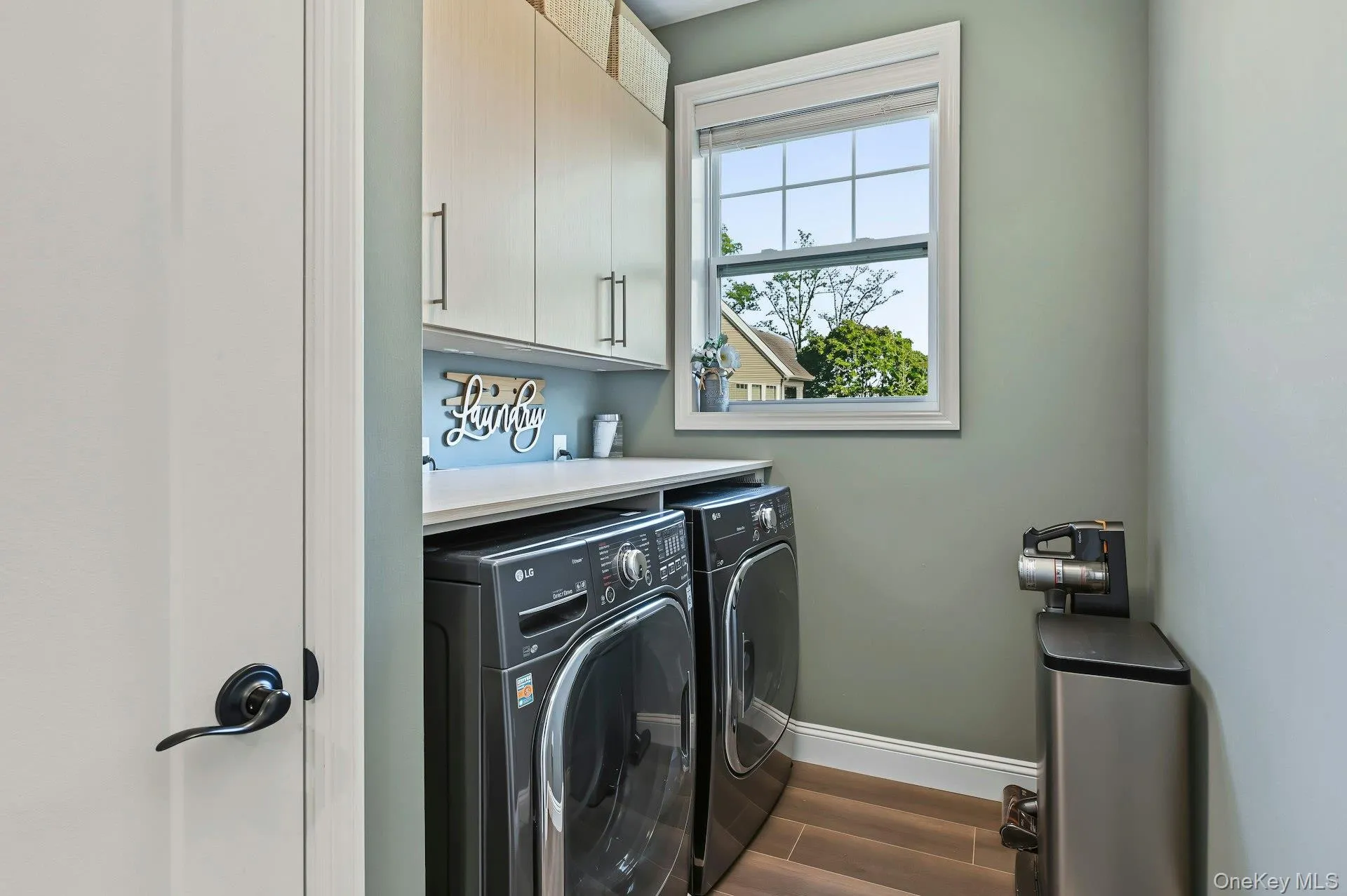Laundry room featuring custom light-finish cabinetry, a solid surface countertop, wood-finish flooring, and a single-hung window with a roller shade Laundry room featuring custom light-finish cabinetry, a solid surface countertop, wood-finish flooring, and a single-hung window with a roller shade