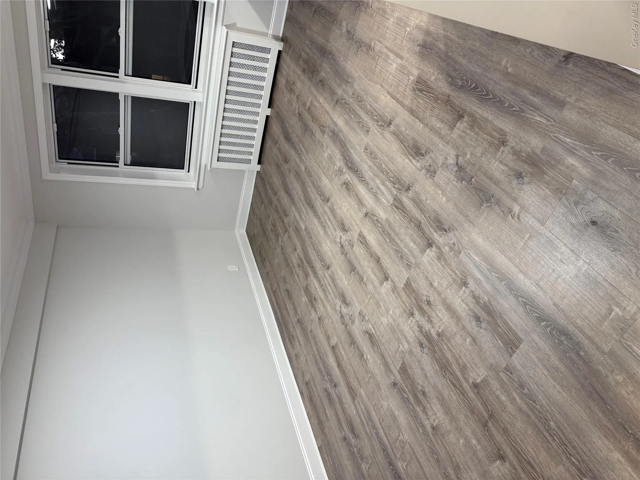 Neutral-toned interior room featuring wood-finish flooring, white window casing, and a built-in radiator cover with decorative lattice screening Neutral-toned interior room featuring wood-finish flooring, white window casing, and a built-in radiator cover with decorative lattice screening