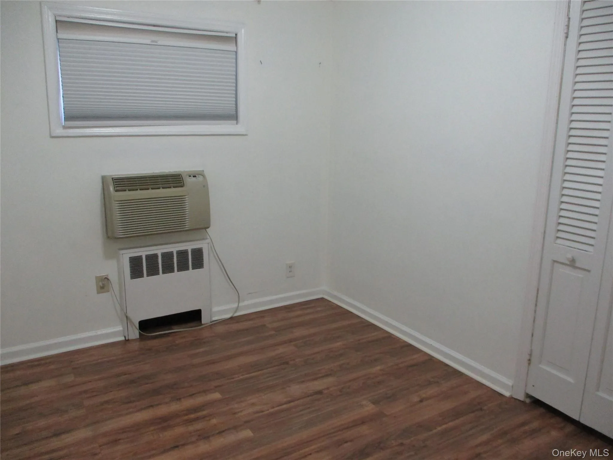 Unfurnished bedroom with radiator heating unit, dark wood-style floors, a wall mounted AC, and a closet Unfurnished bedroom with radiator heating unit, dark wood-style floors, a wall mounted AC, and a closet