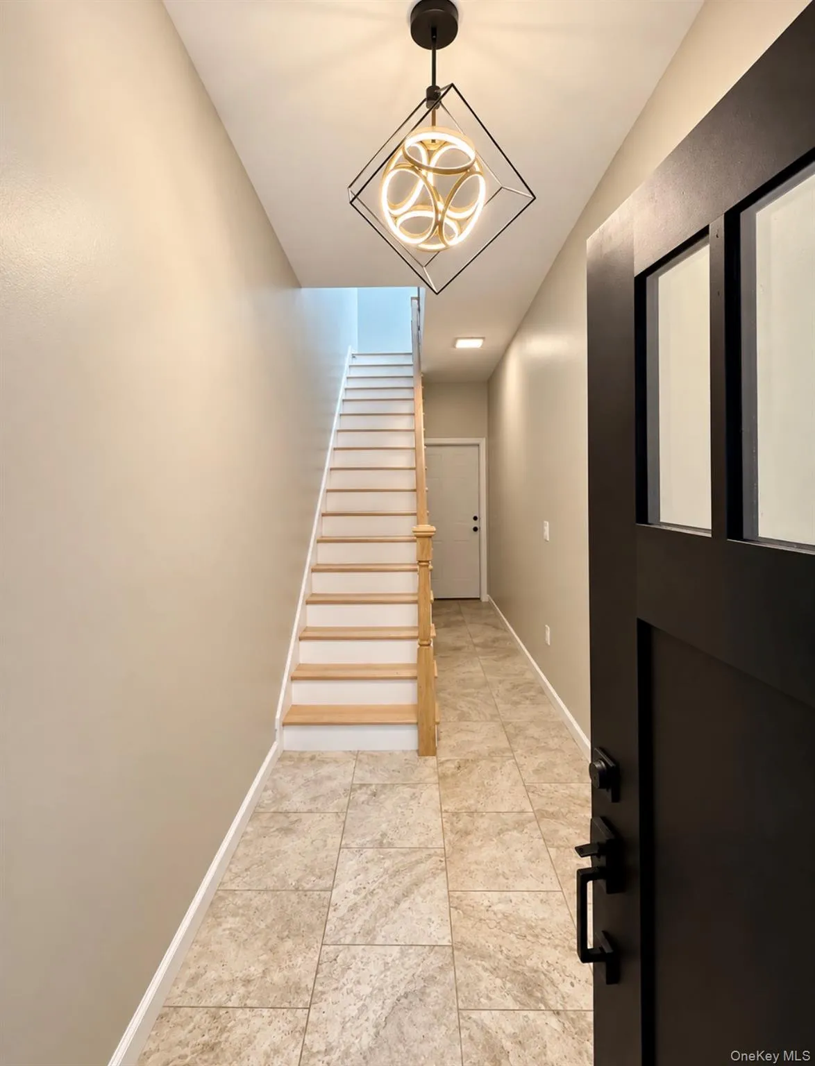 Entryway featuring neutral stone-finish tile flooring, a geometric metal chandelier, a dark-finish door with glass panels, and a staircase with light-tone wood-finish treads and a matching handrail Entryway featuring neutral stone-finish tile flooring, a geometric metal chandelier, a dark-finish door with glass panels, and a staircase with light-tone wood-finish treads and a matching handrail