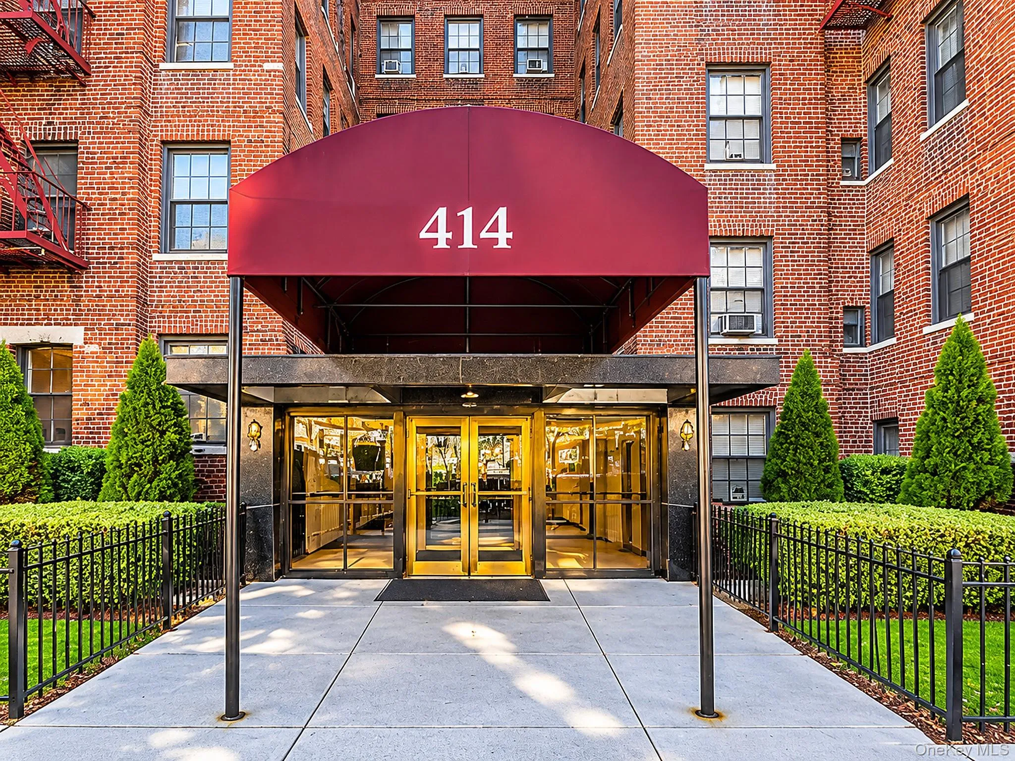 Classic red brick exterior facade featuring a prominent burgundy arched awning, glass-paneled double entry doors with gold-toned framing, and professionally manicured landscaping Classic red brick exterior facade featuring a prominent burgundy arched awning, glass-paneled double entry doors with gold-toned framing, and professionally manicured landscaping