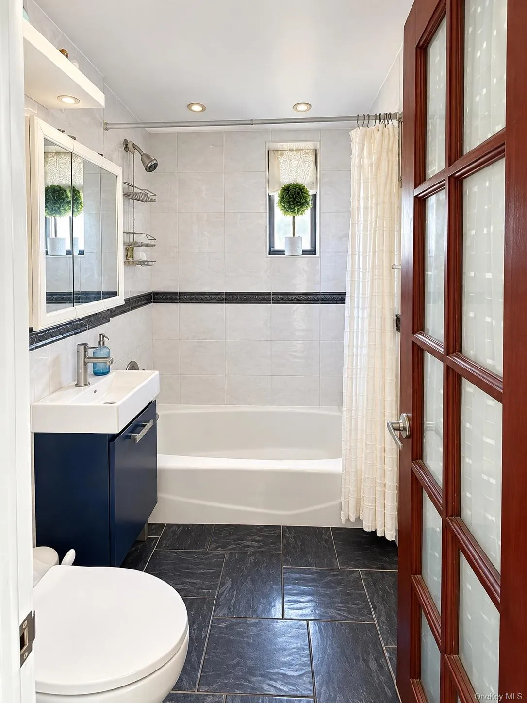 Bathroom featuring slate-finish tile flooring, a wall-mounted vanity with a rectangular basin, recessed ceiling lighting, a soaking tub with tiled surround, and a wood-frame door with frosted glass panels Bathroom featuring slate-finish tile flooring, a wall-mounted vanity with a rectangular basin, recessed ceiling lighting, a soaking tub with tiled surround, and a wood-frame door with frosted glass panels