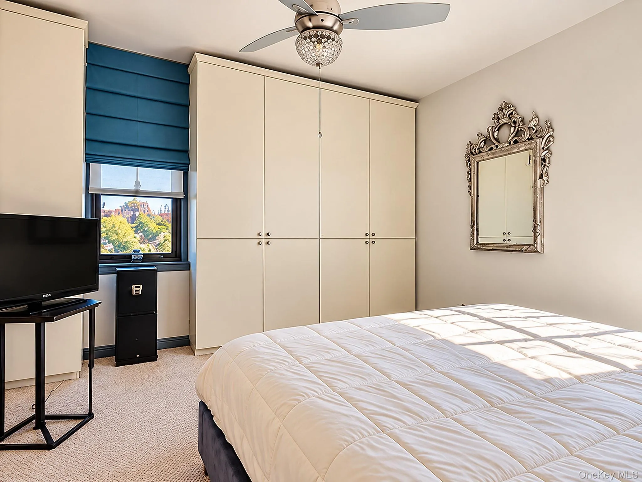 Built-in floor-to-ceiling cabinetry with white finish, integrated ceiling fan with crystal-style light fixture, window with roller shade, and neutral wall-to-wall carpeting Built-in floor-to-ceiling cabinetry with white finish, integrated ceiling fan with crystal-style light fixture, window with roller shade, and neutral wall-to-wall carpeting