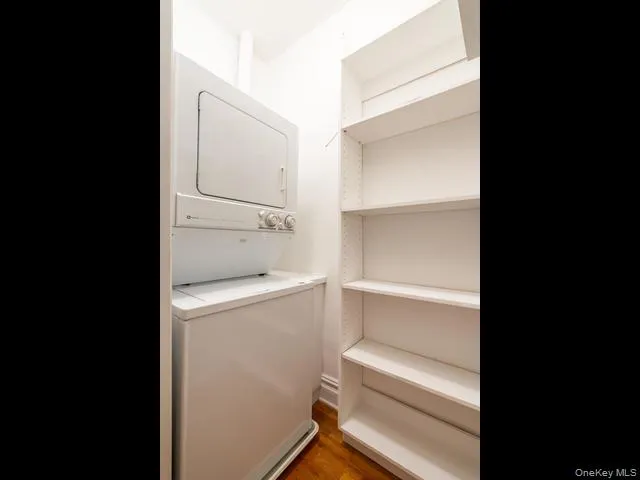 In unit laundry area featuring a full size stackable washer and dryer unit with integrated white built-in shelving and wood-finish flooring In unit laundry area featuring a full size stackable washer and dryer unit with integrated white built-in shelving and wood-finish flooring