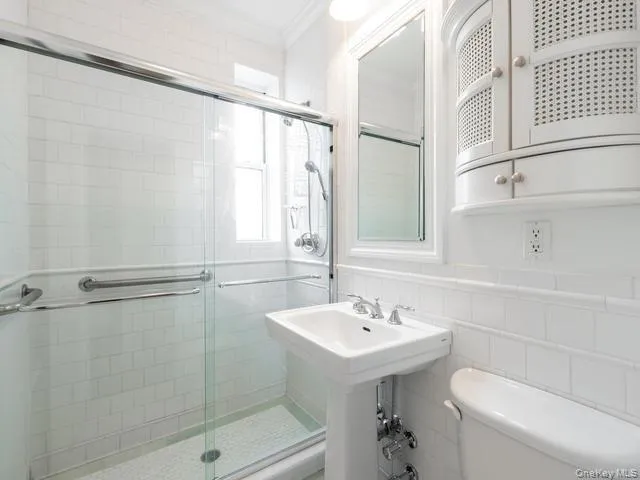 Bathroom featuring a white subway tile surround, glass-enclosed shower, pedestal sink, wall-mounted cabinet with cane-patterned doors, and chrome fixtures Bathroom featuring a white subway tile surround, glass-enclosed shower, pedestal sink, wall-mounted cabinet with cane-patterned doors, and chrome fixtures