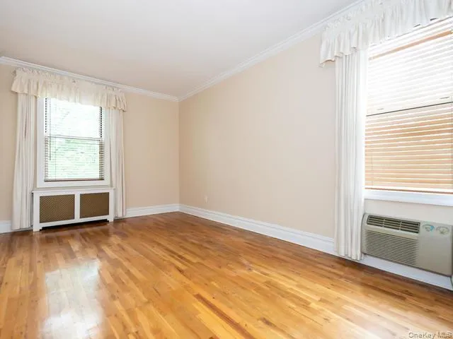 Room featuring wood-finish flooring, crown molding, and two windows with metallic lattice radiator cover and wall-mounted air conditioning unit Room featuring wood-finish flooring, crown molding, and two windows with metallic lattice radiator cover and wall-mounted air conditioning unit