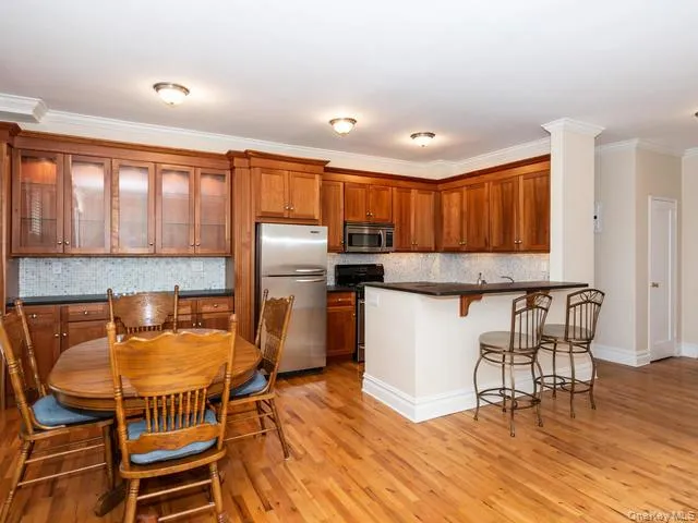 Kitchen featuring wood cabinetry with glass-panel upper doors, stainless steel refrigerator and built-in microwave, granite countertop with breakfast bar, tiled backsplash, and wood-finish flooring Kitchen featuring wood cabinetry with glass-panel upper doors, stainless steel refrigerator and built-in microwave, granite countertop with breakfast bar, tiled backsplash, and wood-finish flooring
