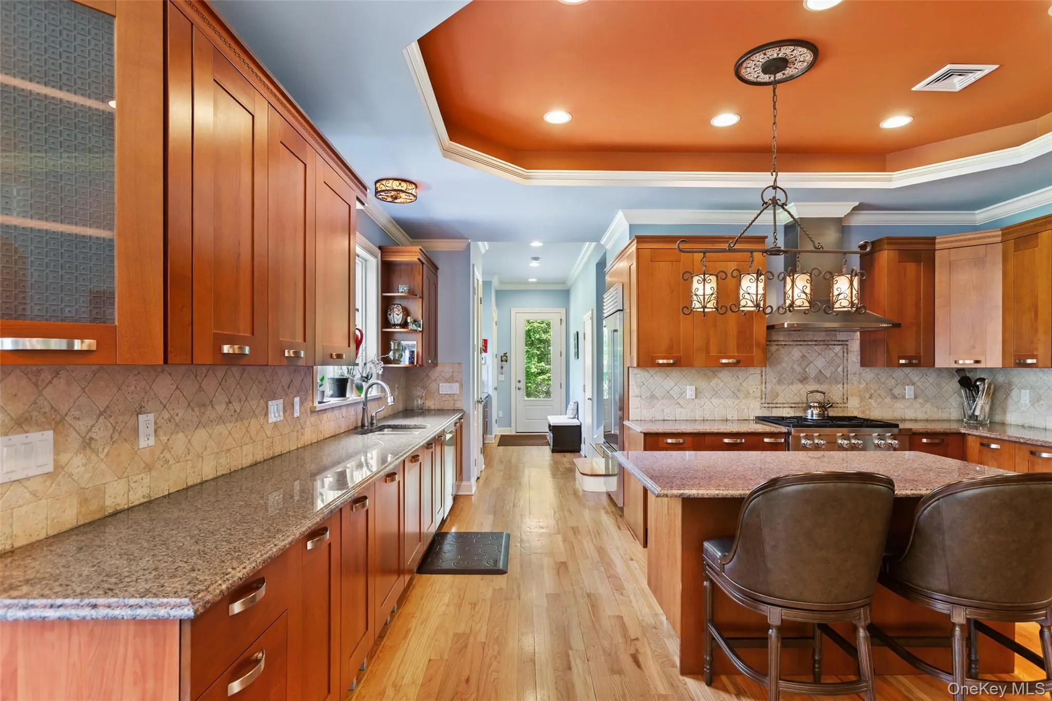 Kitchen with light wood finished floors, a tray ceiling, ornamental molding, wall chimney exhaust hood, and brown cabinets Kitchen with light wood finished floors, a tray ceiling, ornamental molding, wall chimney exhaust hood, and brown cabinets