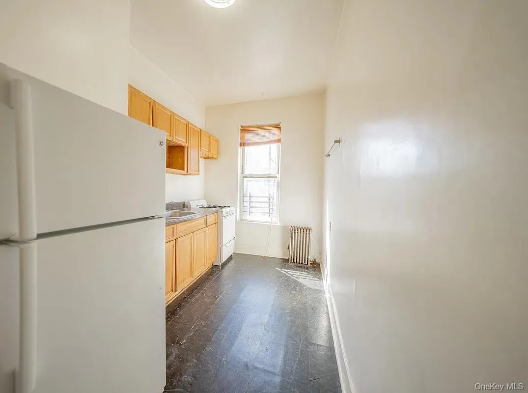 Kitchen featuring wood cabinetry, white appliances, dark flooring, and a window with a roller shade Kitchen featuring wood cabinetry, white appliances, dark flooring, and a window with a roller shade