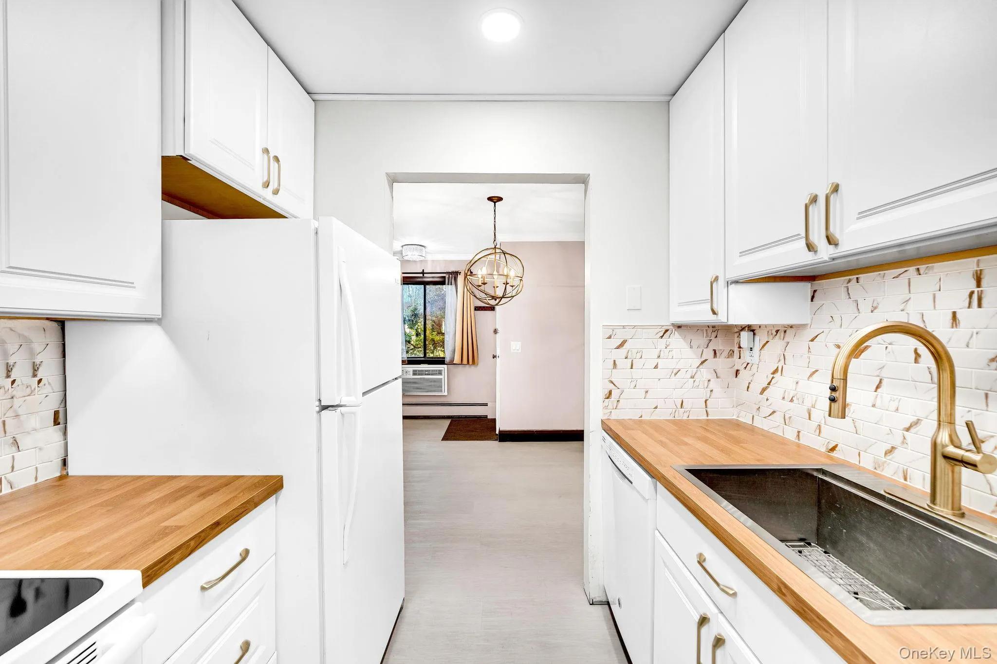 Kitchen featuring white shaker-style cabinetry, butcher block countertops, decorative tile backsplash, undermount sink with a gooseneck faucet, and brass-tone hardware Kitchen featuring white shaker-style cabinetry, butcher block countertops, decorative tile backsplash, undermount sink with a gooseneck faucet, and brass-tone hardware