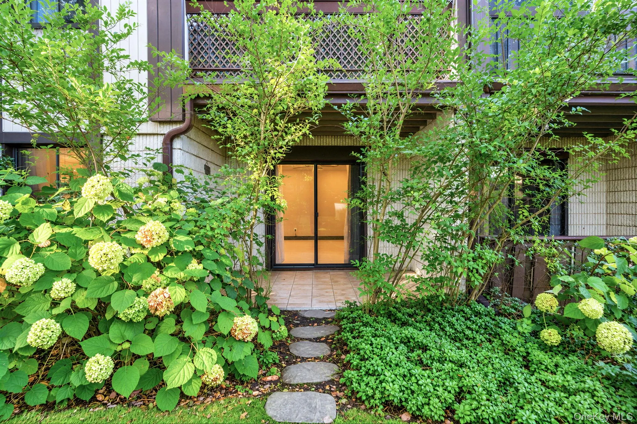 Private patio featuring stone-tiled flooring, a stone paver walkway, mature hydrangeas, and an upper-level balcony with lattice screening Private patio featuring stone-tiled flooring, a stone paver walkway, mature hydrangeas, and an upper-level balcony with lattice screening