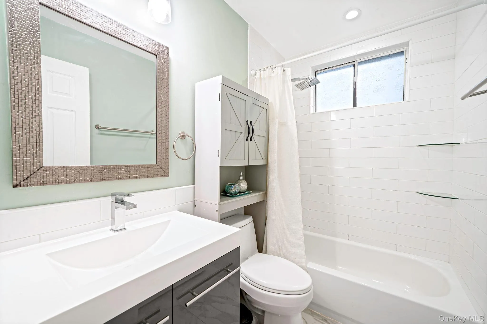 Bathroom featuring a vanity with an integrated basin, a framed wall mirror, subway-tile tub surround, and integrated glass shelving Bathroom featuring a vanity with an integrated basin, a framed wall mirror, subway-tile tub surround, and integrated glass shelving
