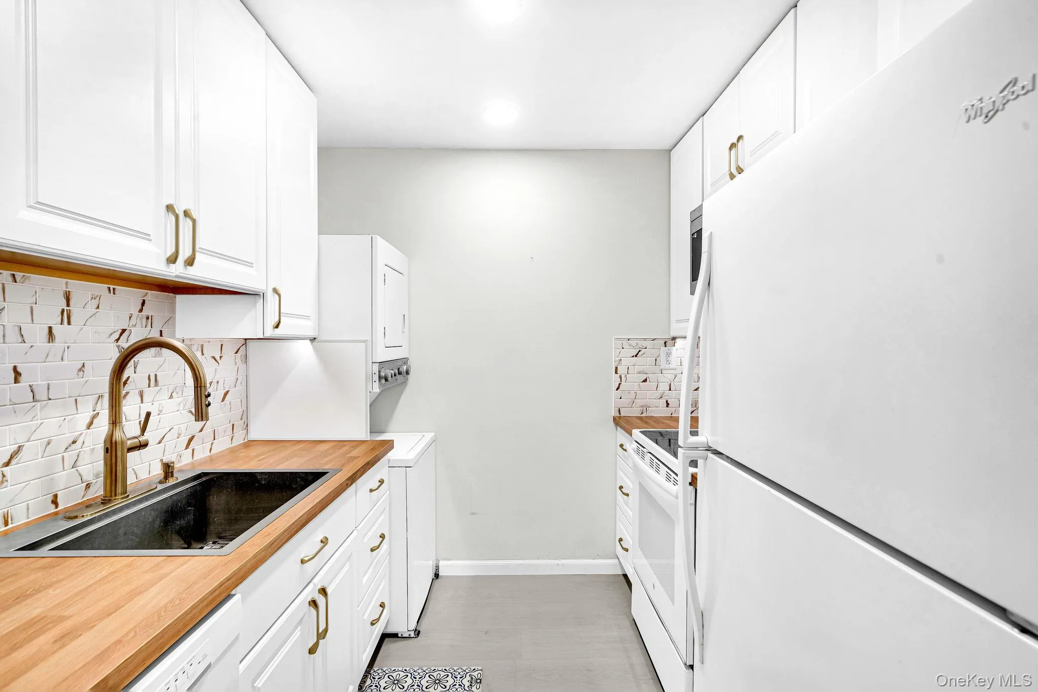 White cabinetry with metallic hardware, butcher block countertops, undermount sink with a gooseneck faucet, tile backsplash, and built-in appliances with washer and dryer in unit. White cabinetry with metallic hardware, butcher block countertops, undermount sink with a gooseneck faucet, tile backsplash, and built-in appliances with washer and dryer in unit.