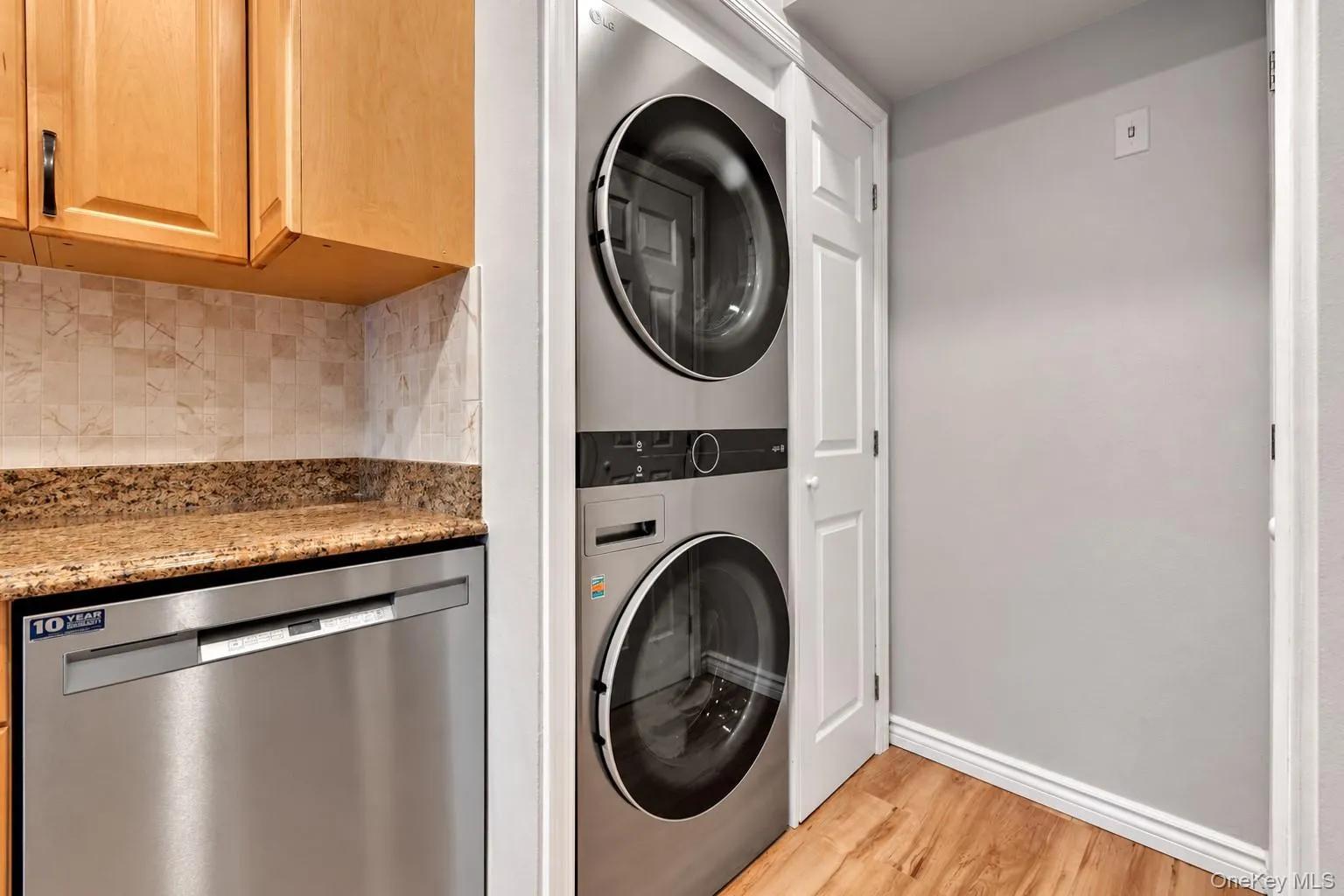 Stacked stainless steel laundry center, wood-finish flooring, stone-finish countertops, tiled backsplash, and built-in wood cabinetry Stacked stainless steel laundry center, wood-finish flooring, stone-finish countertops, tiled backsplash, and built-in wood cabinetry