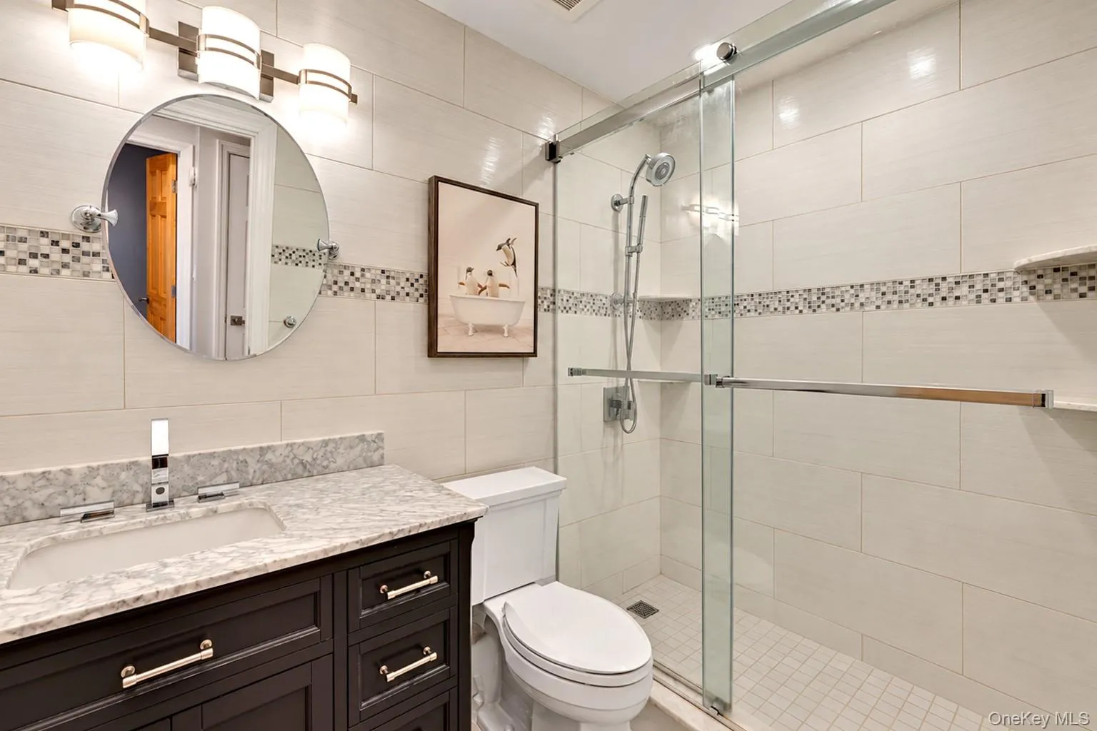 Dark-finish vanity with a stone countertop, circular mirror, multi-light fixture, and walk-in shower with a sliding glass door and mosaic tile inlay Dark-finish vanity with a stone countertop, circular mirror, multi-light fixture, and walk-in shower with a sliding glass door and mosaic tile inlay