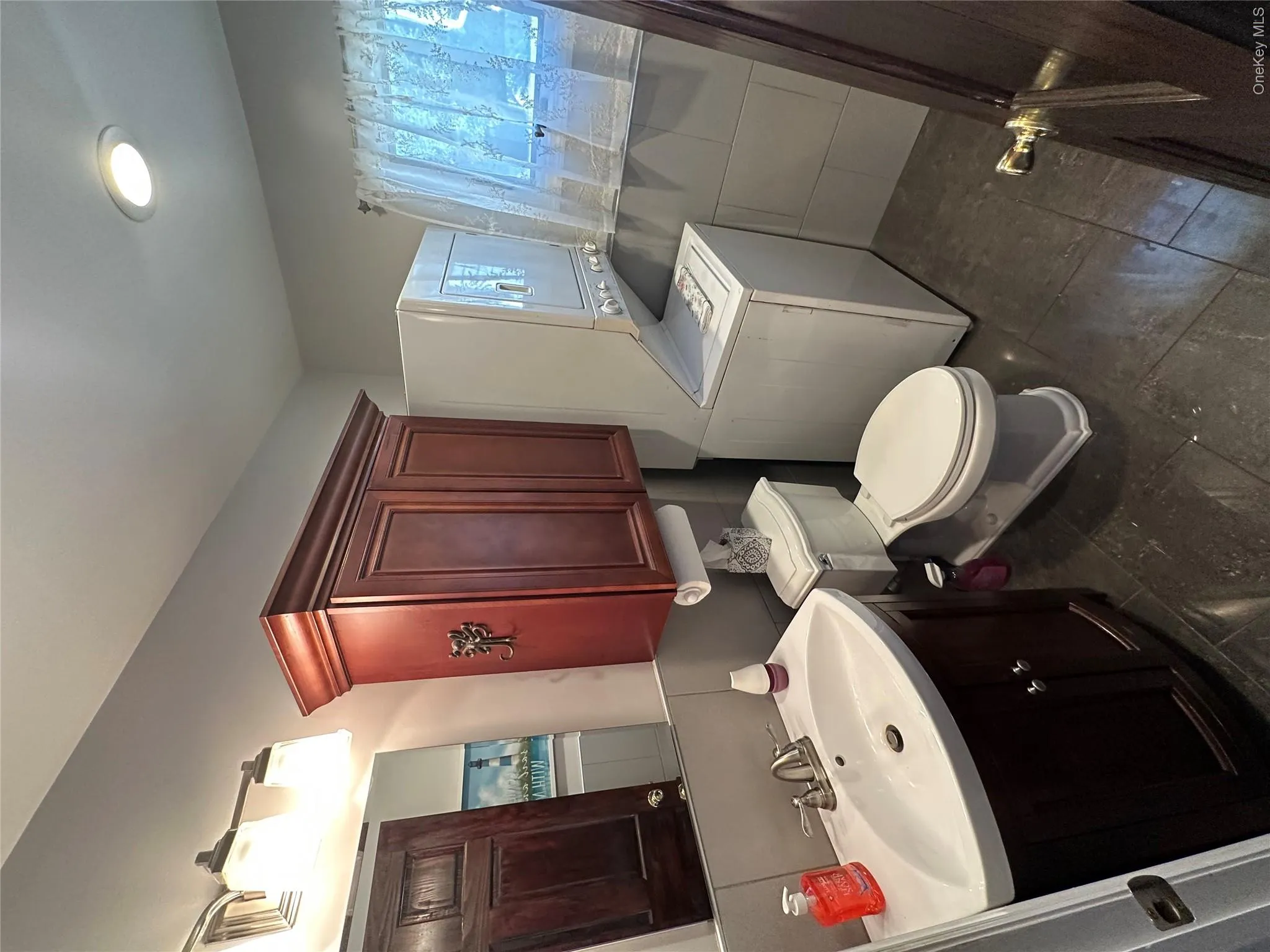 Bathroom featuring dark wood-finish cabinetry, a ceramic basin with a polished chrome faucet, large-format tile flooring, and integrated stacked laundry appliances Bathroom featuring dark wood-finish cabinetry, a ceramic basin with a polished chrome faucet, large-format tile flooring, and integrated stacked laundry appliances