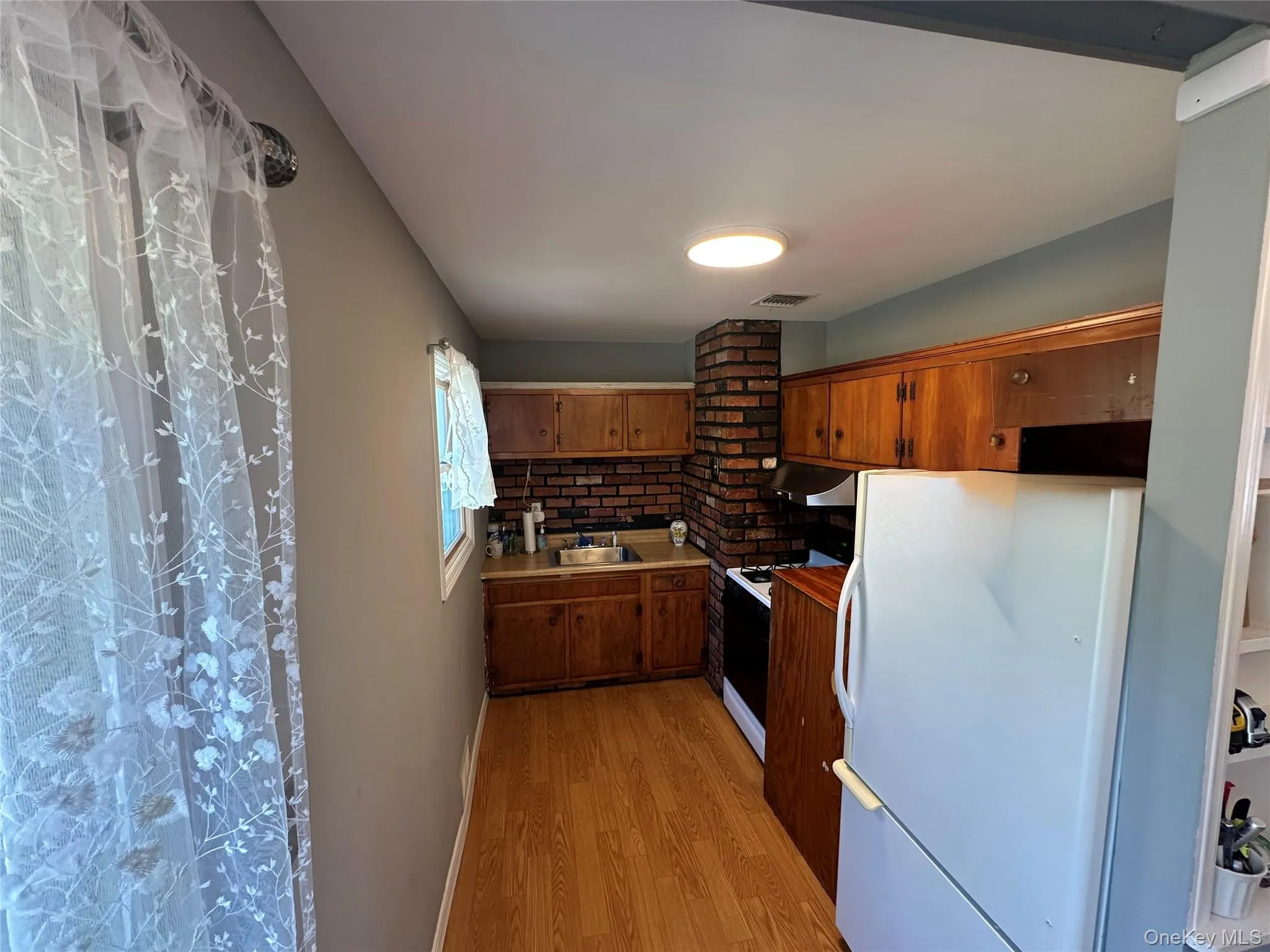 Kitchen featuring wood cabinetry, brick accent wall, wood-finish flooring, and built-in flush-mount lighting Kitchen featuring wood cabinetry, brick accent wall, wood-finish flooring, and built-in flush-mount lighting