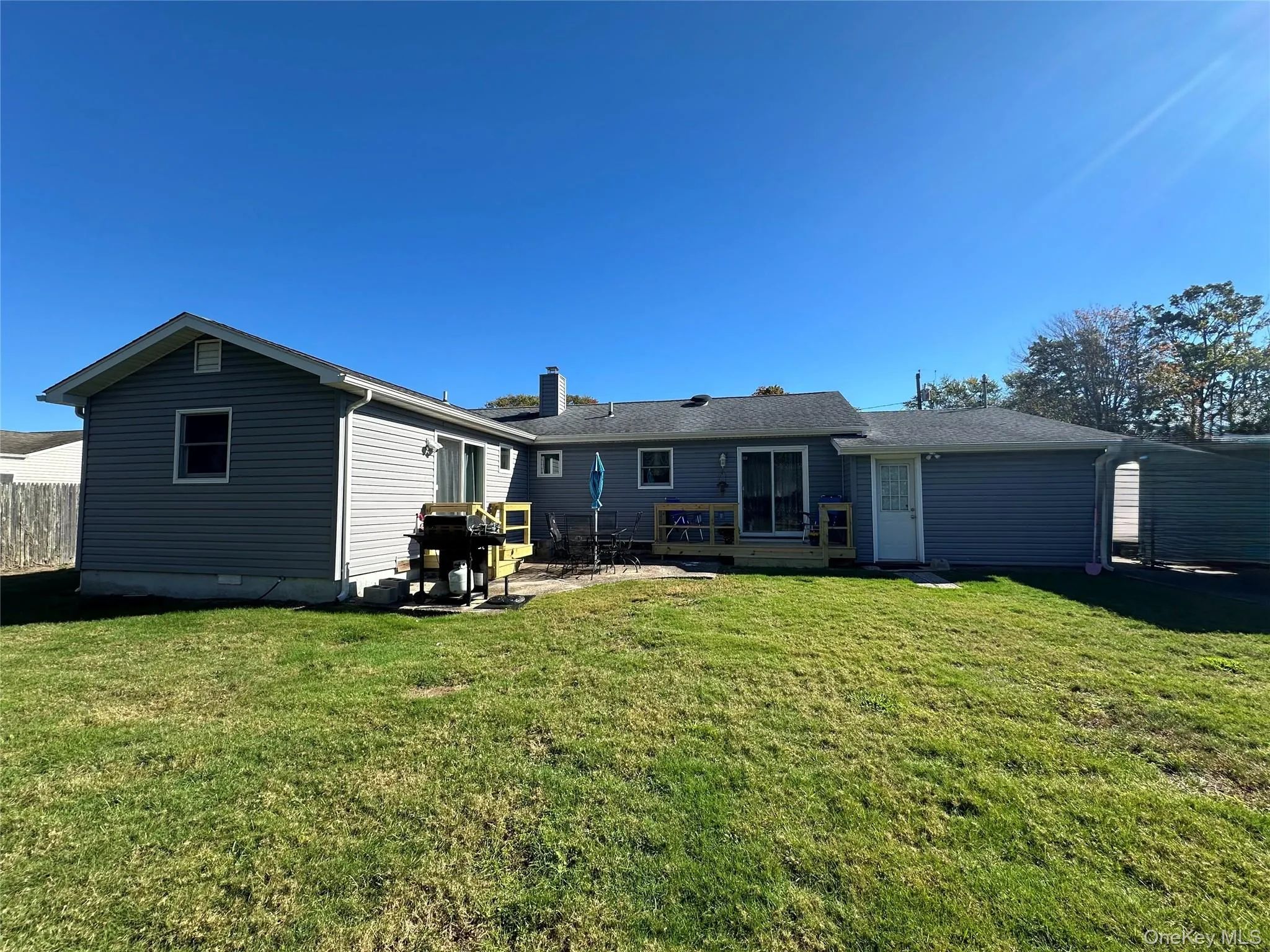 Rear exterior featuring light grey vinyl siding, a paved patio area, wood decking, and a partially fenced backyard Rear exterior featuring light grey vinyl siding, a paved patio area, wood decking, and a partially fenced backyard