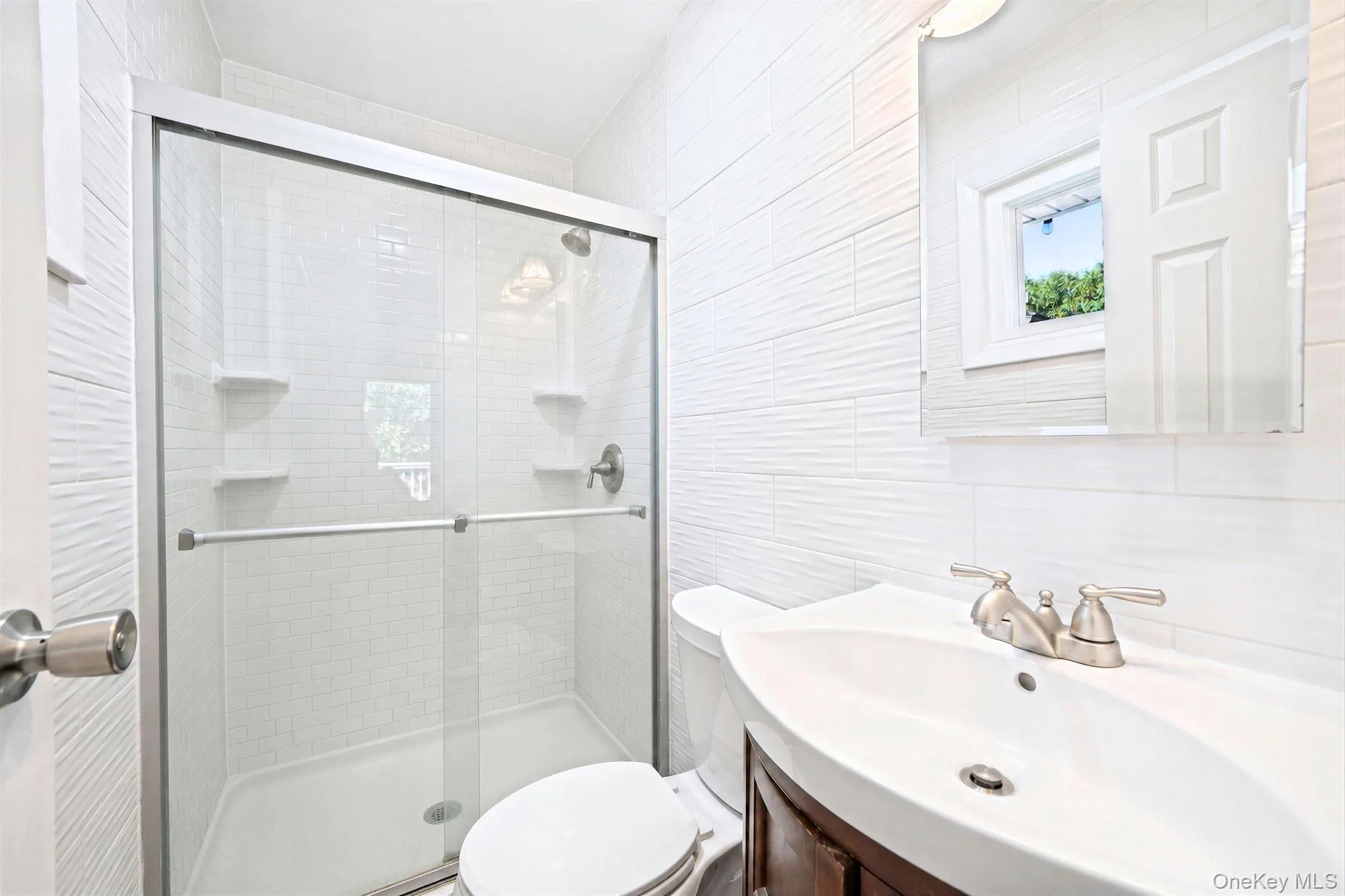Bathroom featuring white textured wall tile, a glass-enclosed shower with built-in shelving, an integrated vanity sink, and a wall-mounted mirror Bathroom featuring white textured wall tile, a glass-enclosed shower with built-in shelving, an integrated vanity sink, and a wall-mounted mirror