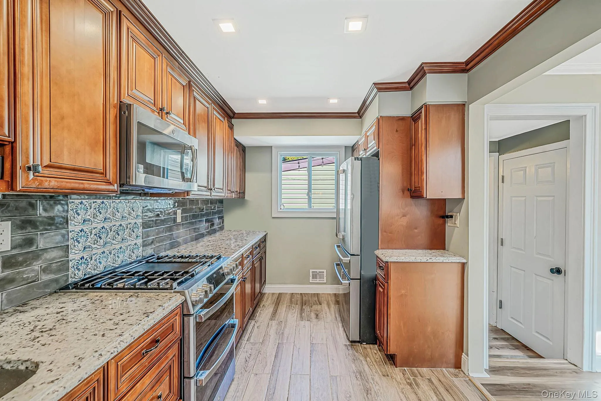 Kitchen featuring wood cabinetry, stone-finish countertops, stainless steel appliances, decorative tile backsplash, and wood-finish flooring Kitchen featuring wood cabinetry, stone-finish countertops, stainless steel appliances, decorative tile backsplash, and wood-finish flooring