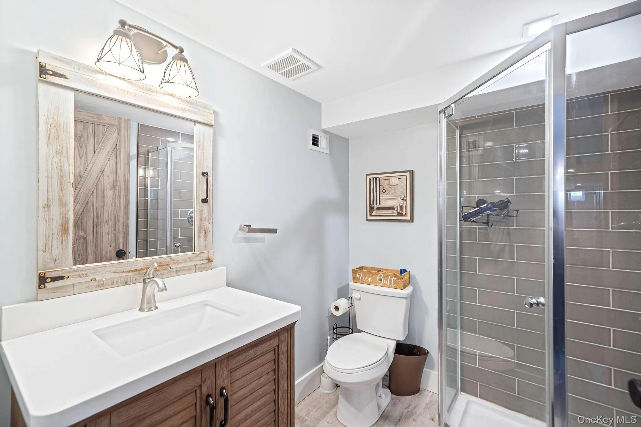 Bathroom featuring a wooden vanity with a white countertop, wood-framed wall mirror, corner glass-enclosed shower with tiled walls, and metallic lighting fixtures Bathroom featuring a wooden vanity with a white countertop, wood-framed wall mirror, corner glass-enclosed shower with tiled walls, and metallic lighting fixtures