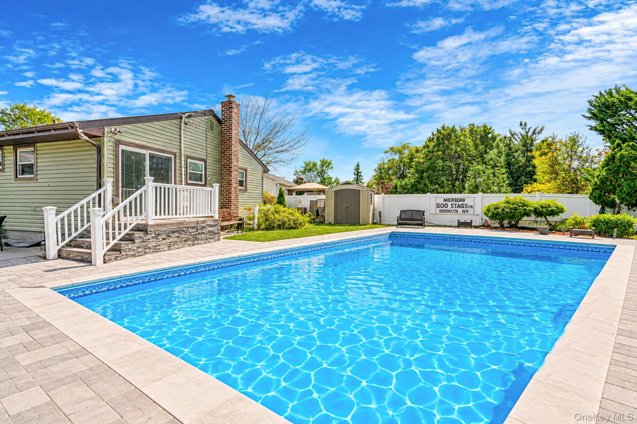 In-ground swimming pool with stone-style paver decking, white vinyl privacy fencing, raised deck with white railings, and a brick chimney exterior In-ground swimming pool with stone-style paver decking, white vinyl privacy fencing, raised deck with white railings, and a brick chimney exterior