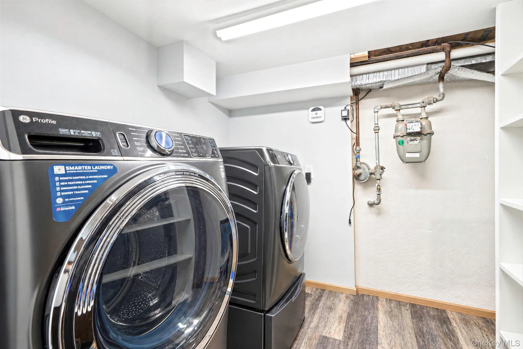 Laundry area featuring wood-finish flooring, white shelving, and dual front-loading appliances Laundry area featuring wood-finish flooring, white shelving, and dual front-loading appliances