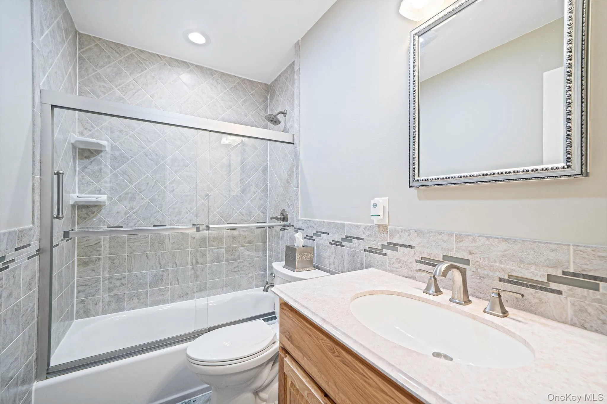 Bathroom featuring a vanity with a light-toned countertop, integrated basin, and brushed nickel finish faucet, complemented by a tiled shower surround with decorative glass mosaic inlay and a sliding glass door Bathroom featuring a vanity with a light-toned countertop, integrated basin, and brushed nickel finish faucet, complemented by a tiled shower surround with decorative glass mosaic inlay and a sliding glass door