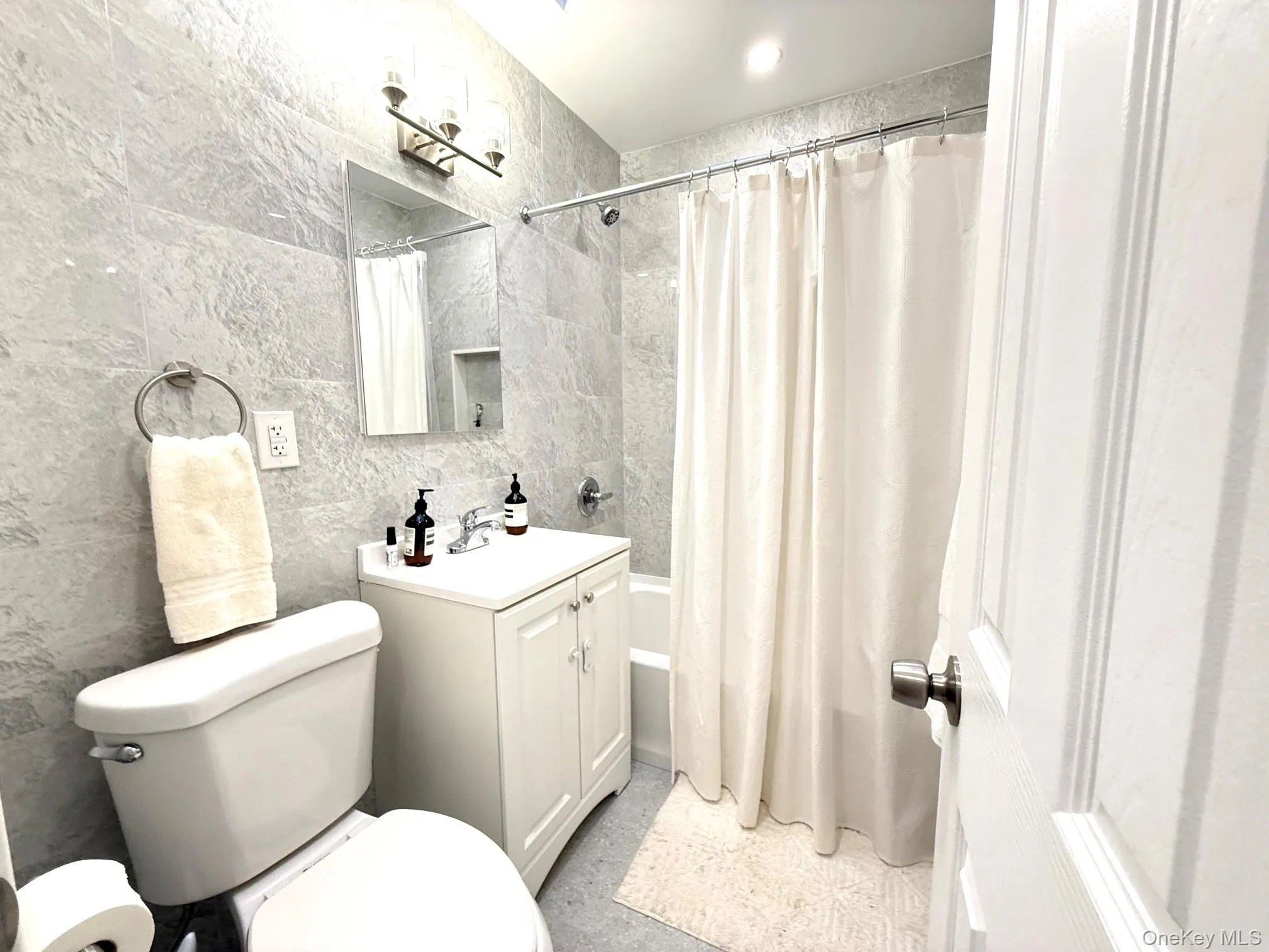 Bathroom featuring floor-to-ceiling stone-textured wall tile, vanity with white cabinetry, rectangular mirror, three-light fixture, and tub-shower combination Bathroom featuring floor-to-ceiling stone-textured wall tile, vanity with white cabinetry, rectangular mirror, three-light fixture, and tub-shower combination