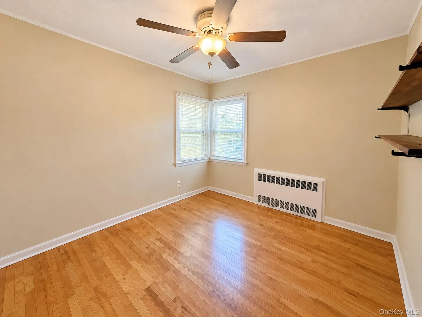 Room featuring wood-finish flooring, a ceiling fan with integrated lighting, corner windows, and built-in wall shelving Room featuring wood-finish flooring, a ceiling fan with integrated lighting, corner windows, and built-in wall shelving