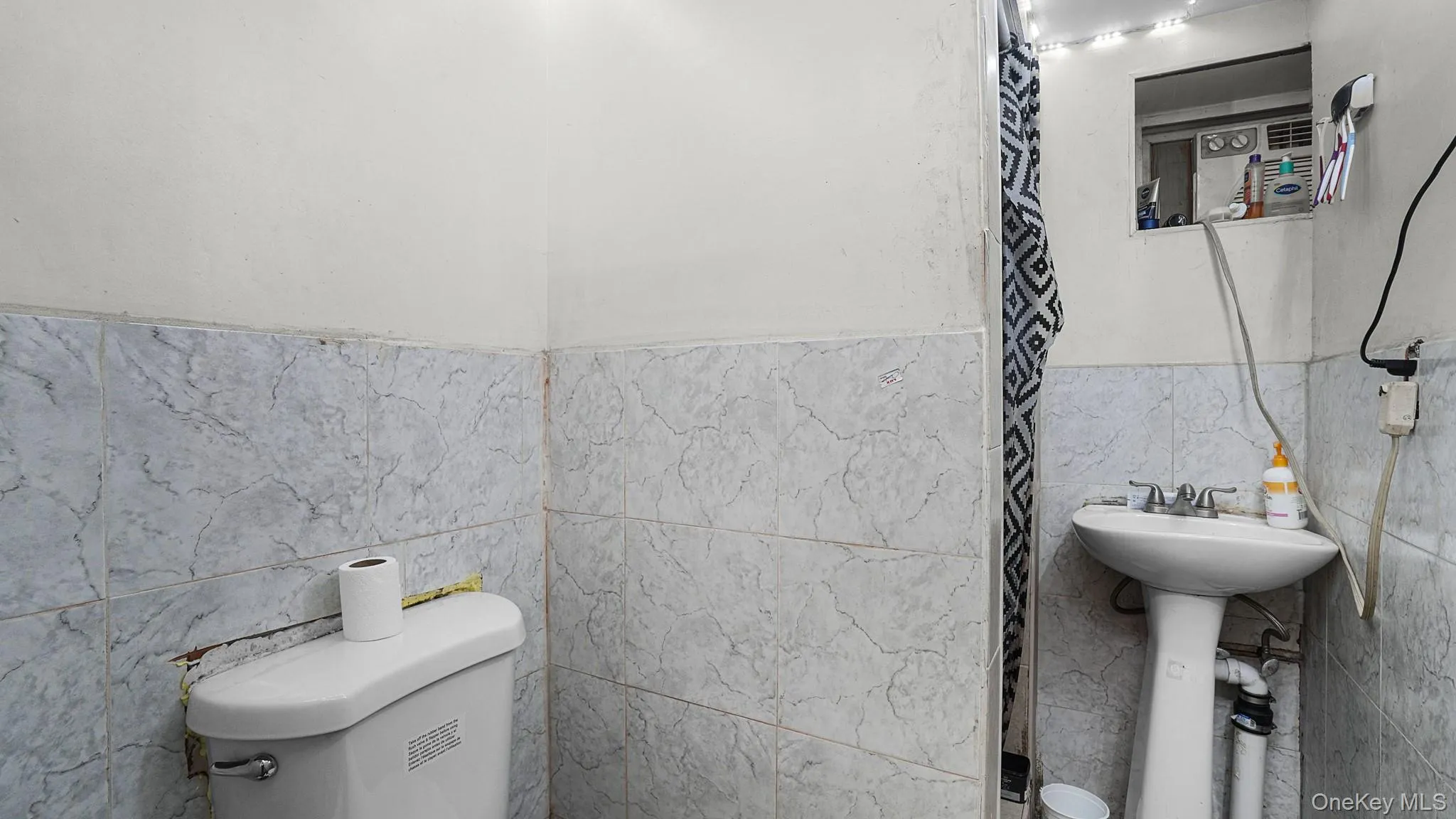 Bathroom featuring wall-to-wall stone-patterned tile, white pedestal sink with dual-handle faucet, and standard porcelain toilet Bathroom featuring wall-to-wall stone-patterned tile, white pedestal sink with dual-handle faucet, and standard porcelain toilet