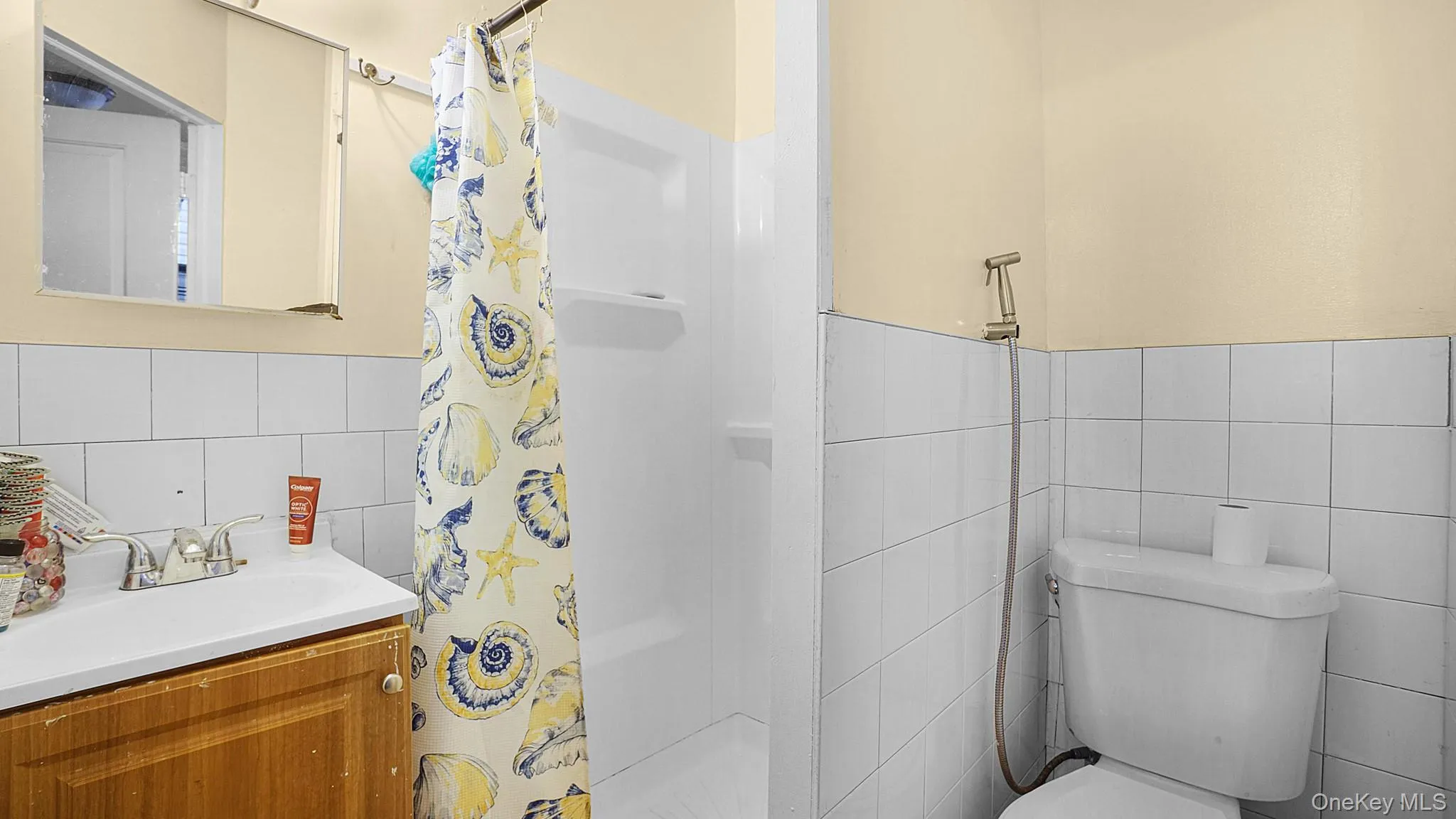Bathroom featuring a wood-finish vanity, white square wall tiling, a white sink with chrome hardware, a shower enclosure with built-in shelving, and a toilet with an attached handheld sprayer Bathroom featuring a wood-finish vanity, white square wall tiling, a white sink with chrome hardware, a shower enclosure with built-in shelving, and a toilet with an attached handheld sprayer