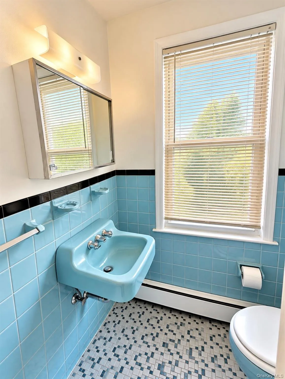 Wall-mounted ceramic basin, medicine cabinet with integrated vanity lighting, dual-tone square tile wainscoting with black accent border, and mosaic tile flooring Wall-mounted ceramic basin, medicine cabinet with integrated vanity lighting, dual-tone square tile wainscoting with black accent border, and mosaic tile flooring